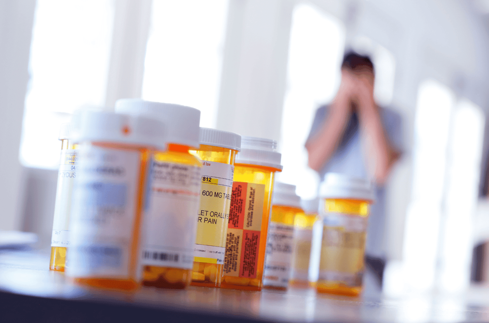 Several prescription pill bottles on a table with a person blurred in the background.