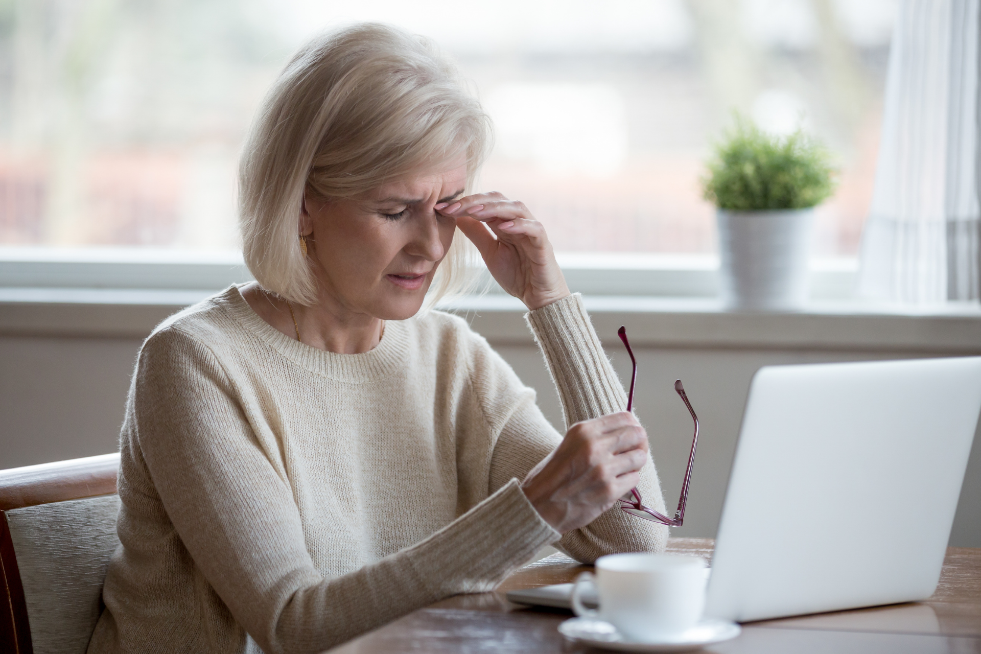 Middle-aged woman rubbing her forehead while holding glasses, looking fatigued as she sits at a table with a laptop and coffee