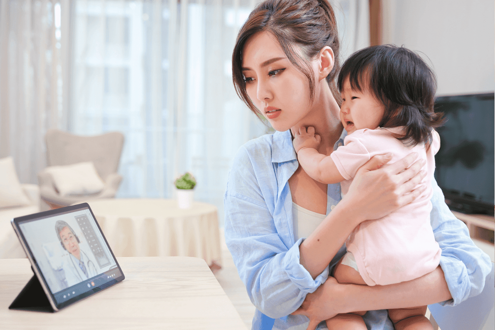 Mother holding a crying baby while having a virtual consultation with a doctor on a tablet.