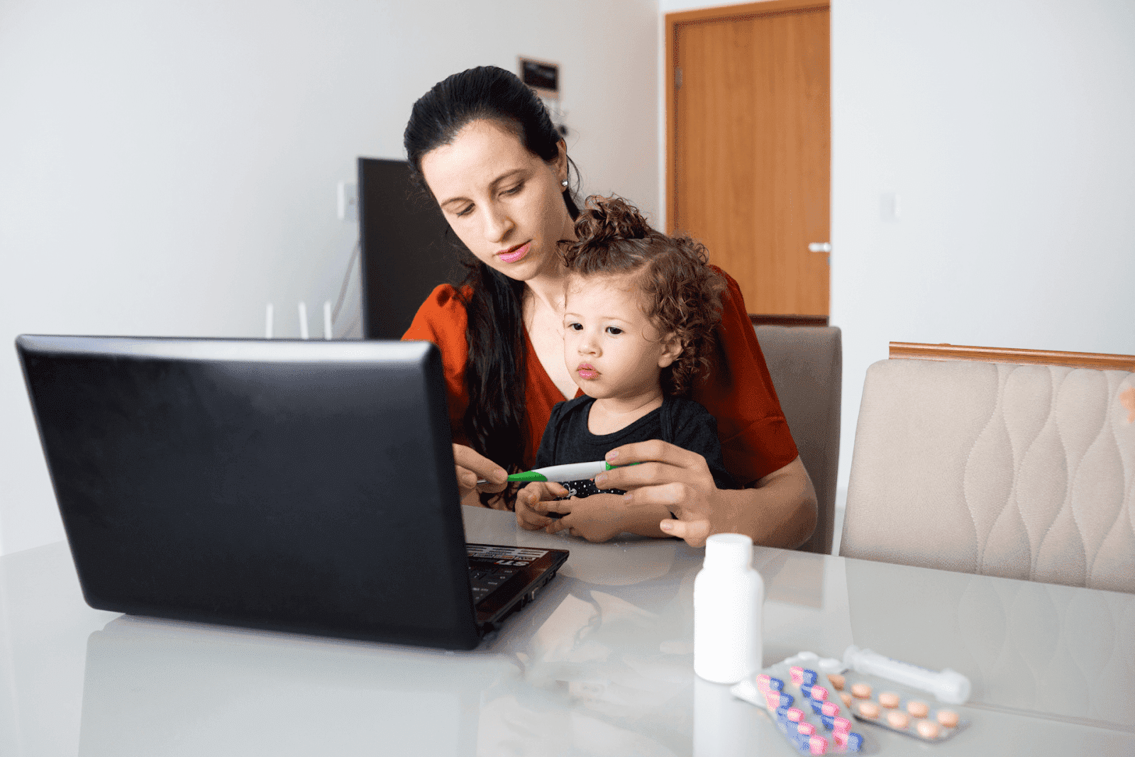 Mother holding a thermometer while sitting with her young child in front of a laptop, with medicine and pills on the table