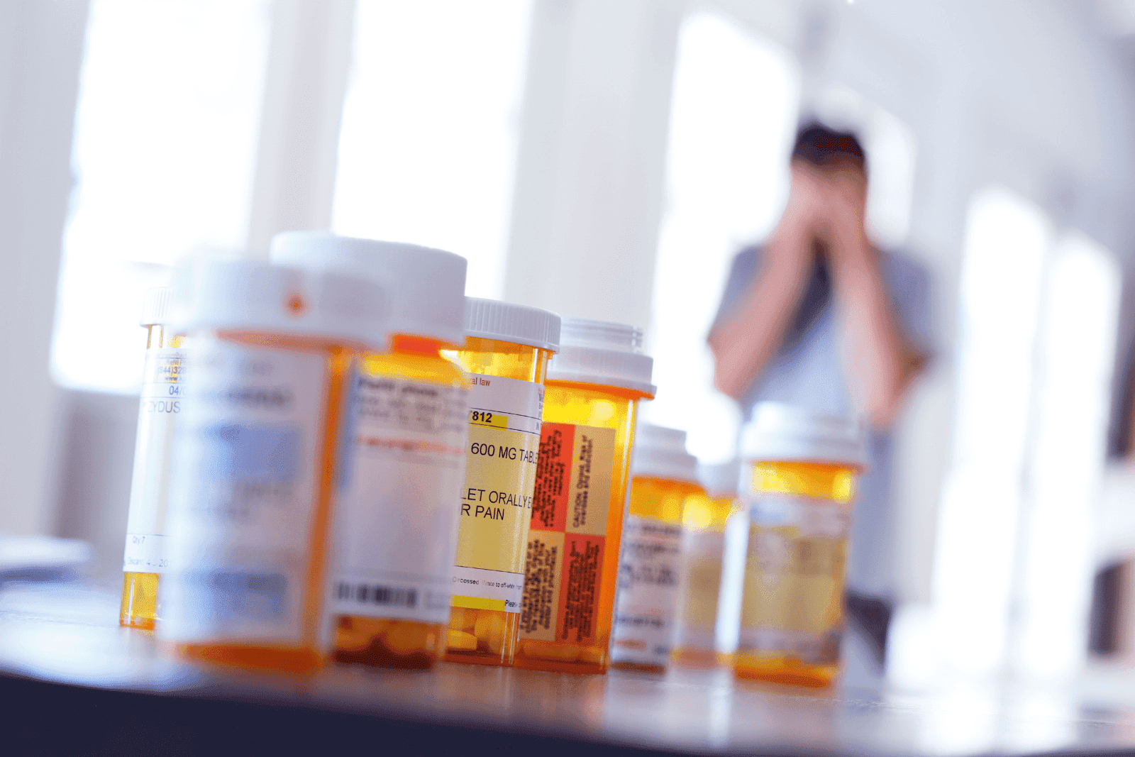 Multiple prescription pill bottles on a table, with a blurred person holding their head in the background