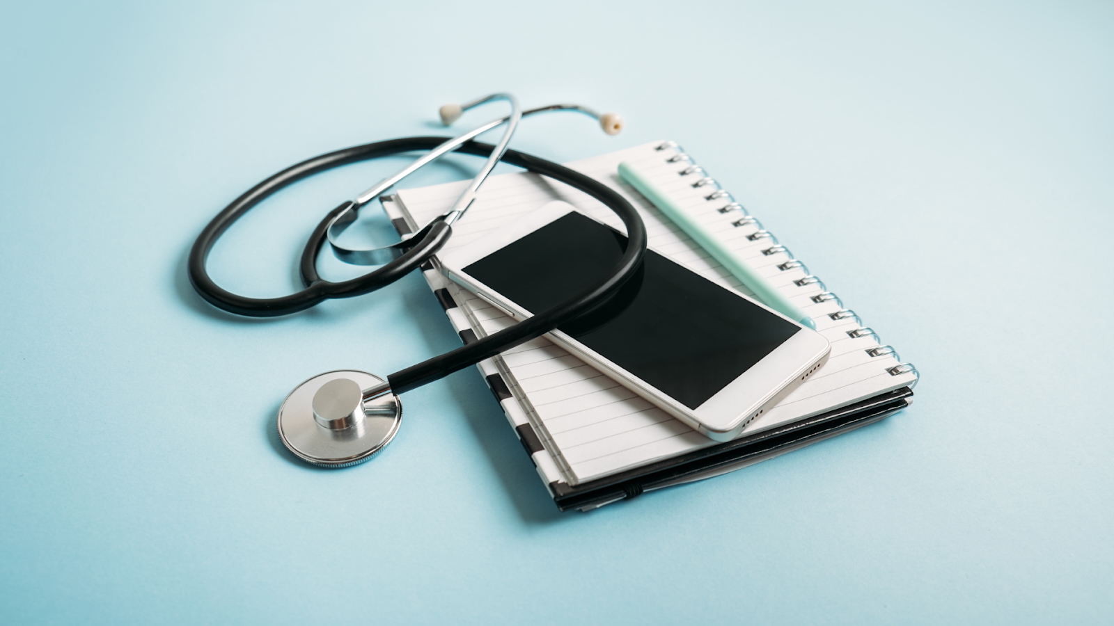 A stethoscope and smartphone rest on a spiral notebook with a pen, symbolizing modern healthcare tools and telemedicine