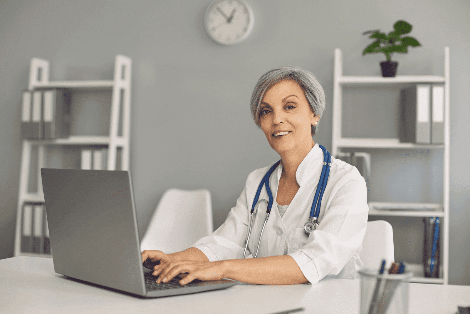 Older female doctor smiling while working on a laptop in her office