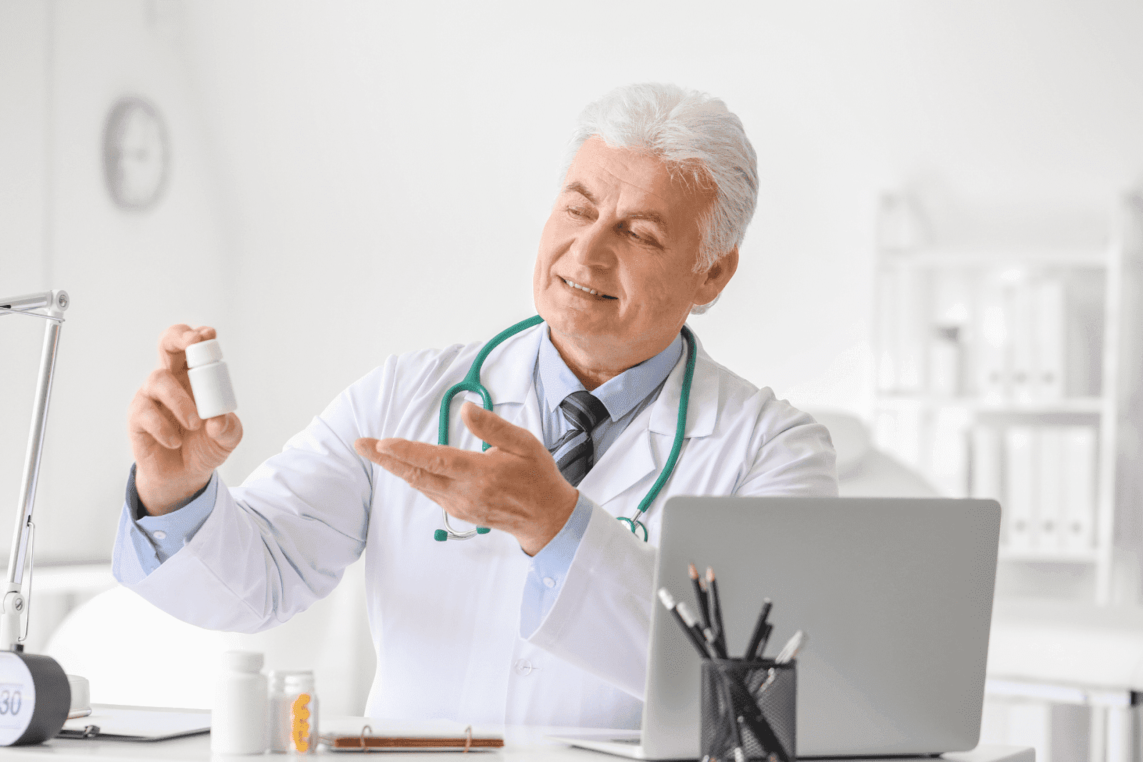 Older male doctor in a white coat holding a pill bottle while sitting at a desk with a laptop