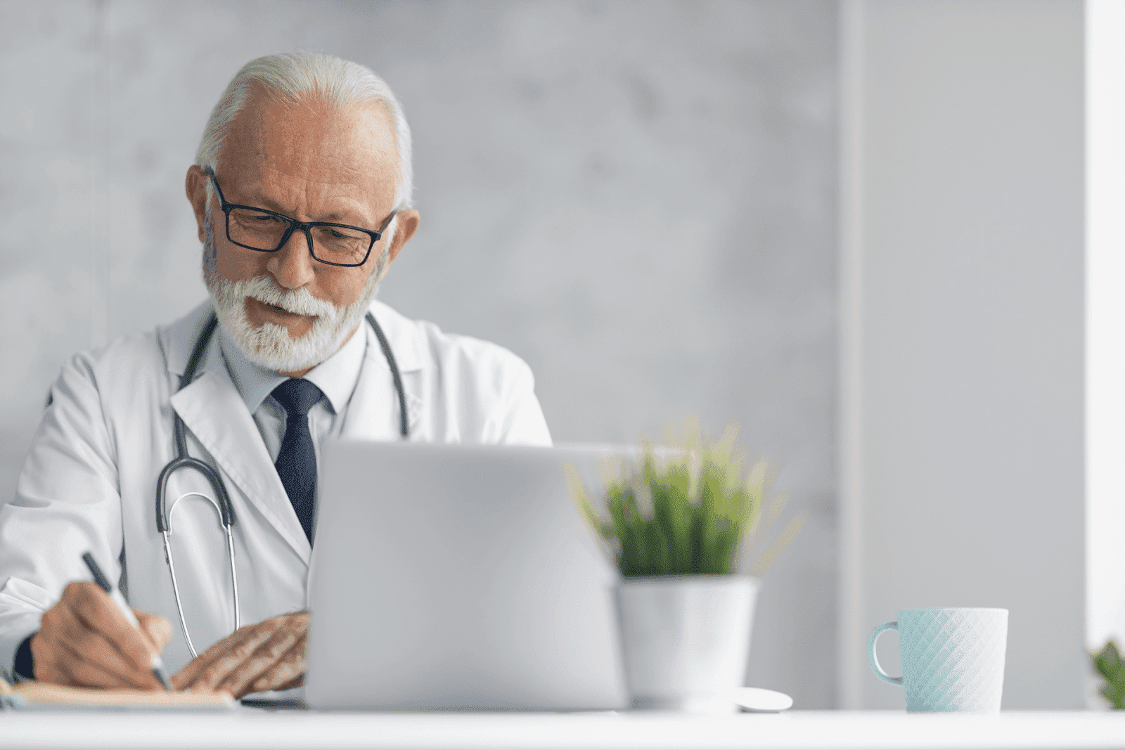 Older male doctor taking notes while working on a laptop at his desk.