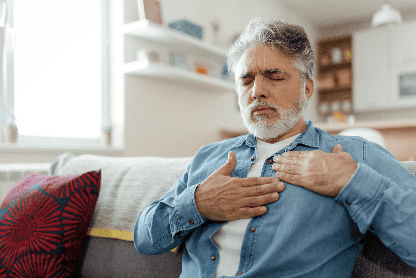 Older man sitting on a couch with hands on his chest, appearing to experience chest pain or heartburn