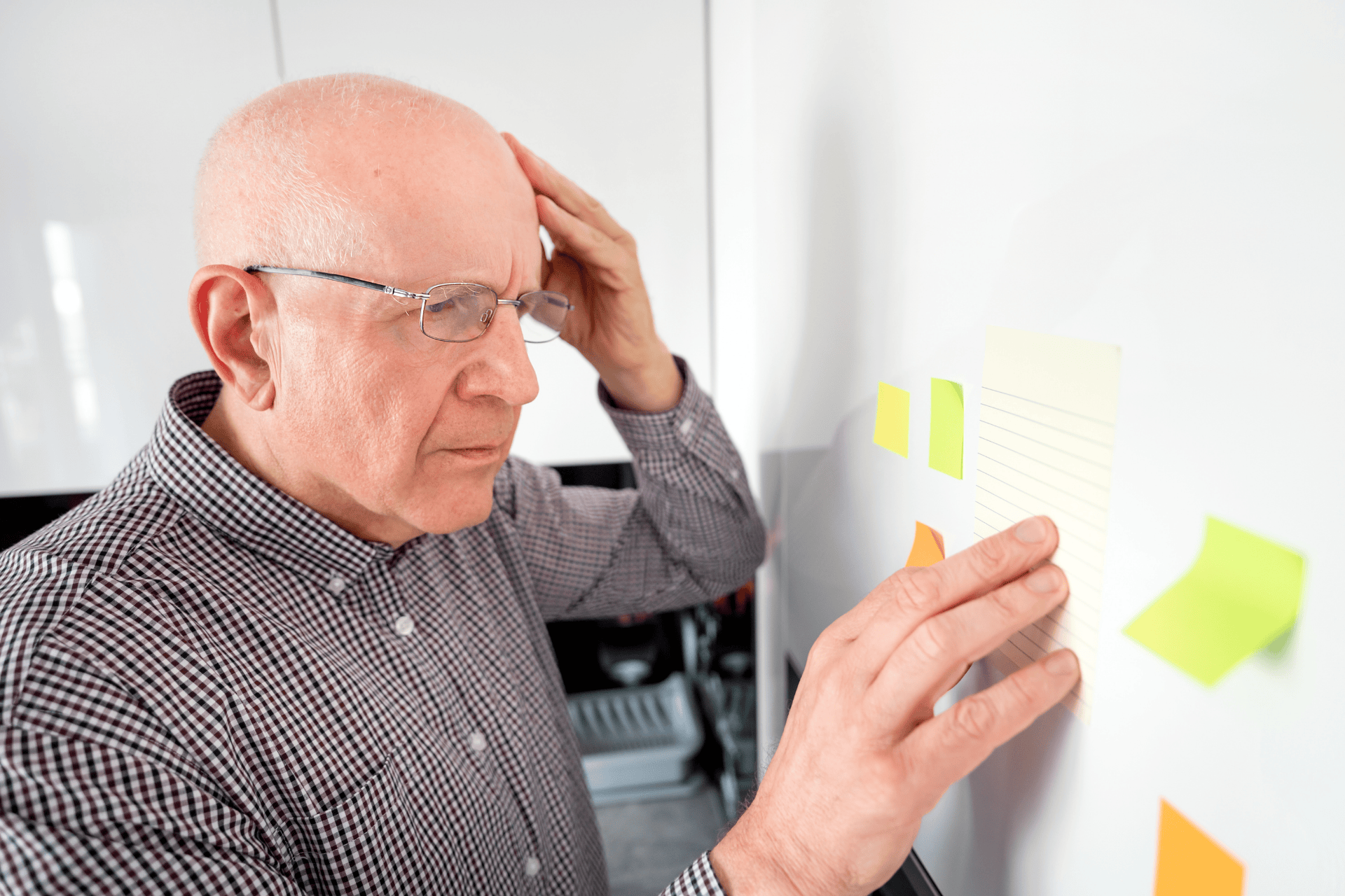 Older man touching his head while looking at sticky notes on a whiteboard.