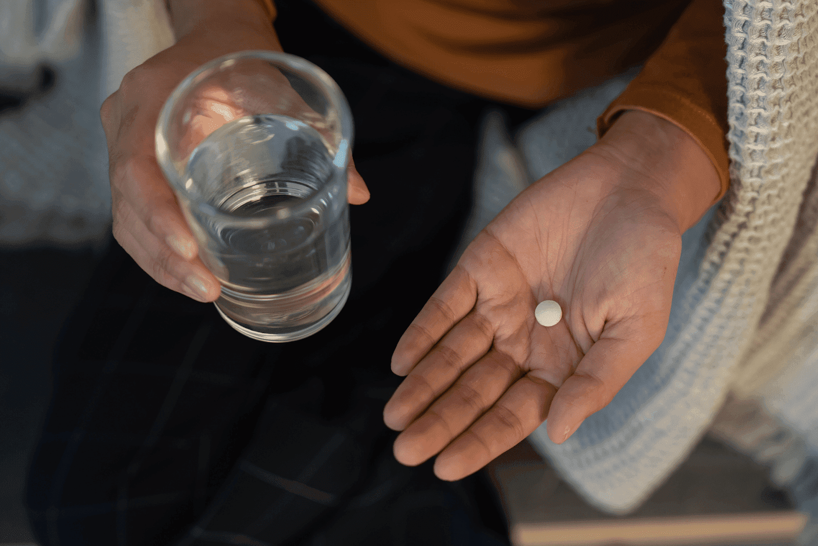 Person holding a glass of water in one hand and a white pill in the other.