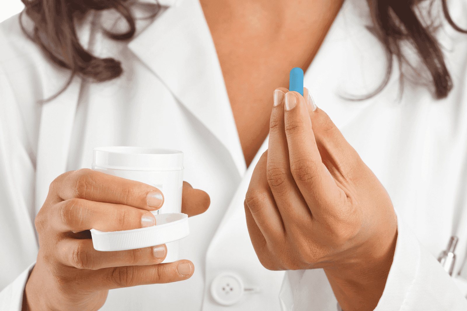 Person in a white coat holding a blue pill in one hand and a white container in the other.