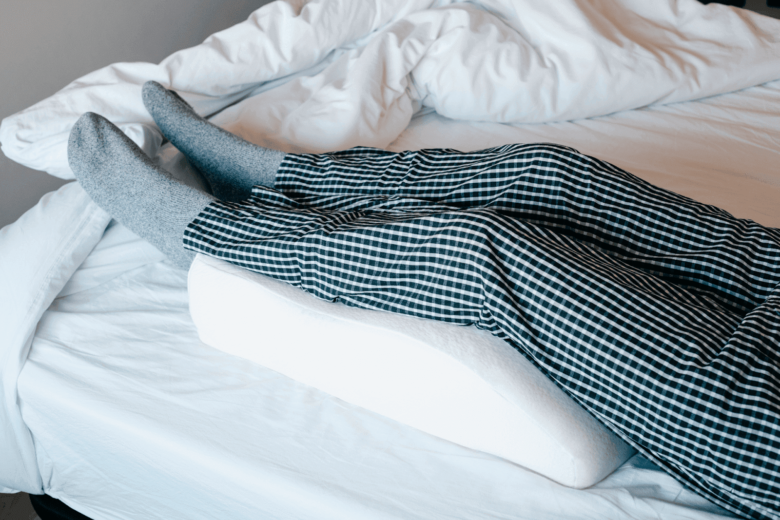 Person lying in bed with legs elevated on a wedge pillow, wearing socks and checkered pajamas