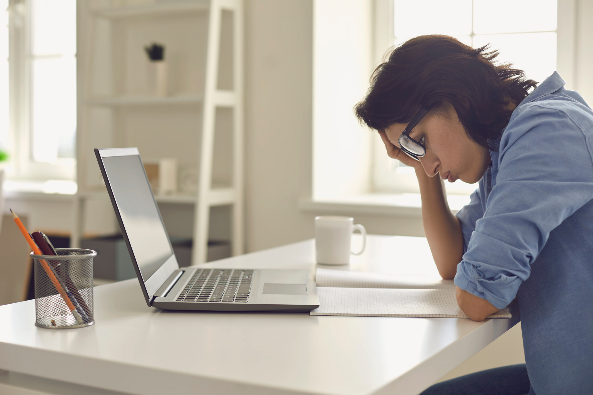 Person sitting at a desk with a laptop, head resting on a hand, looking tired while reading notes