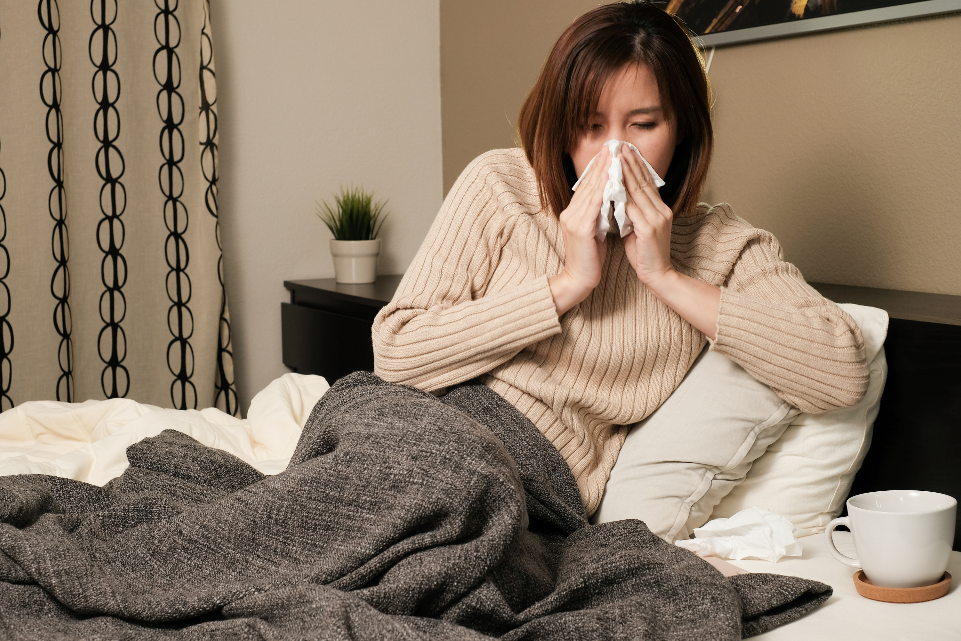 Person sitting up in bed, blowing their nose with a tissue while wrapped in a blanket