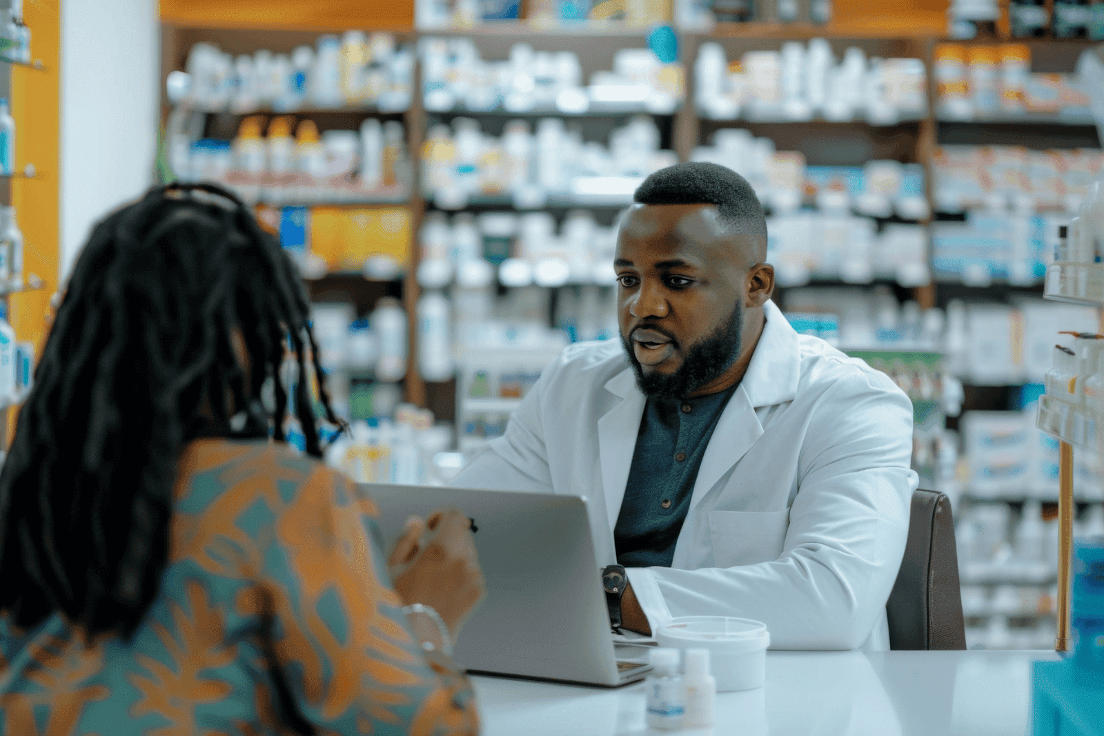 Pharmacist wearing a white coat consulting with a patient across a counter in a pharmacy
