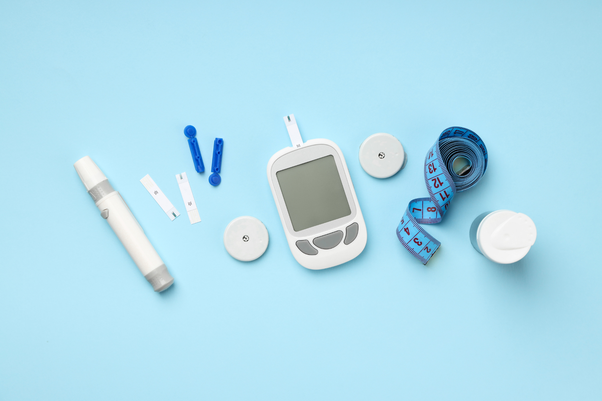 A diabetes testing kit on a blue background: a glucose meter with test strip, lancet device and lancets, test strips, control caps, a roll of measuring tape, and a small bottle