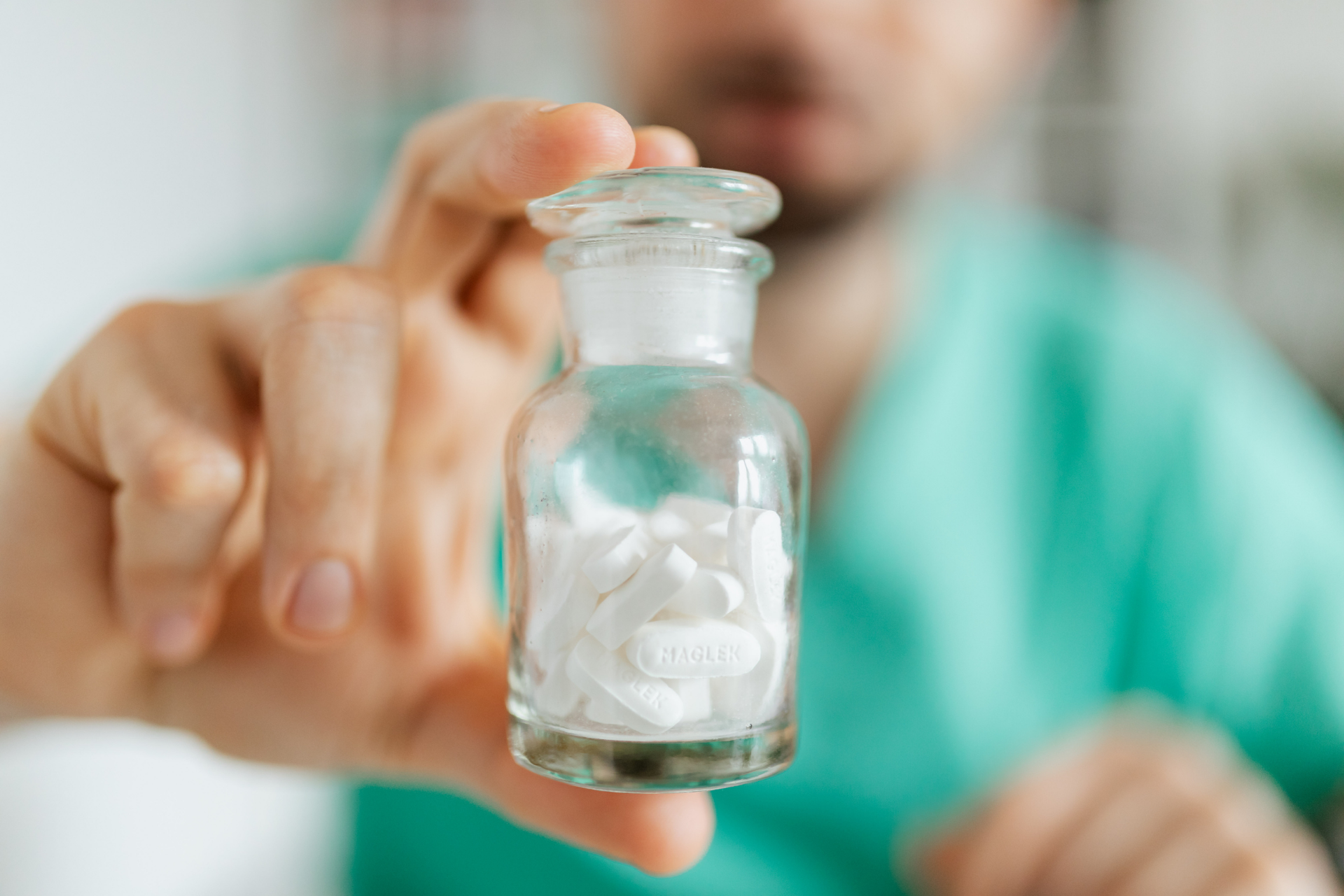 A small glass apothecary jar filled with white tablets, held by a person wearing green scrubs