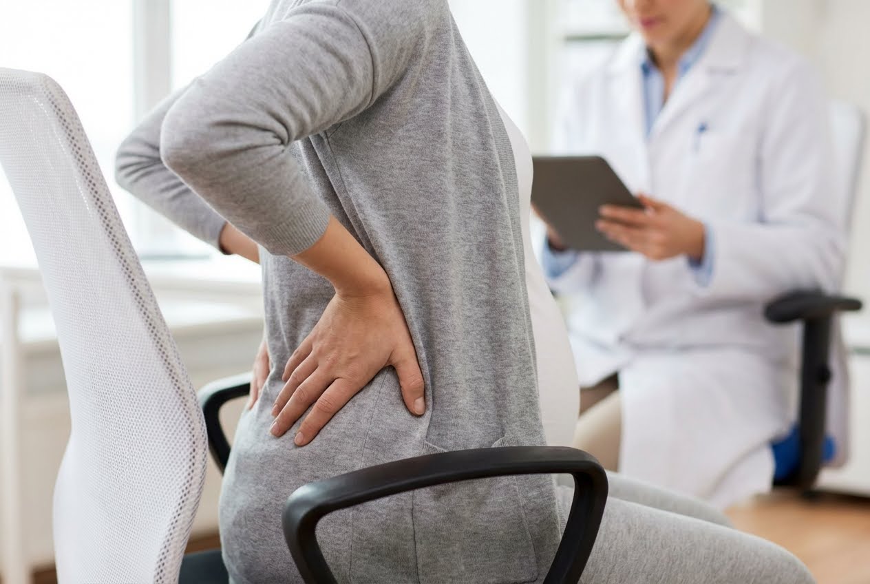 Pregnant woman in gray cardigan holding her lower back in pain, with a doctor in a white coat holding a tablet in the background.