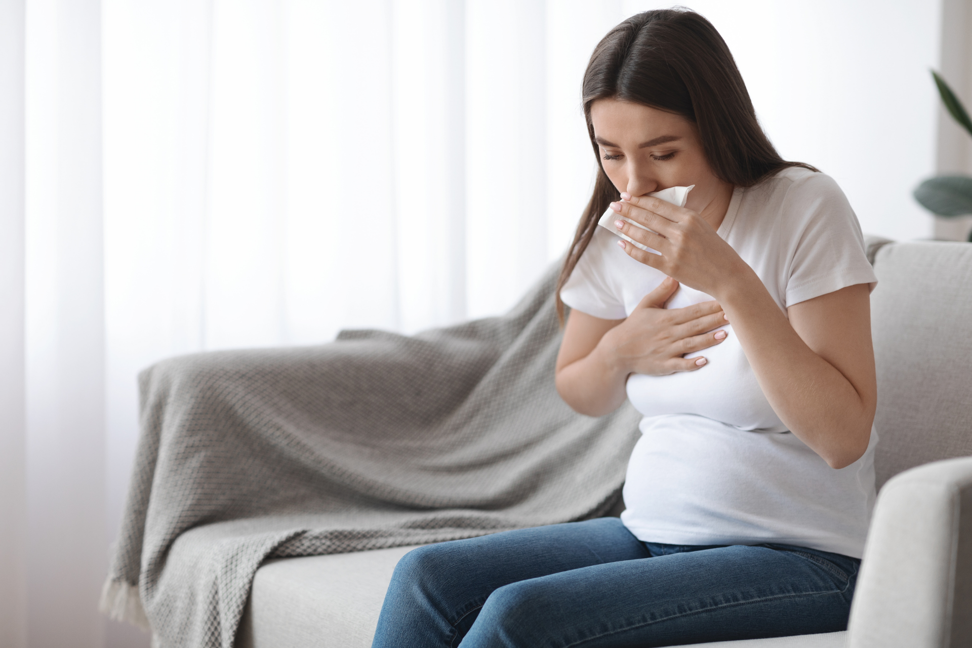 Pregnant woman sitting on a couch, coughing into a tissue while holding her chest