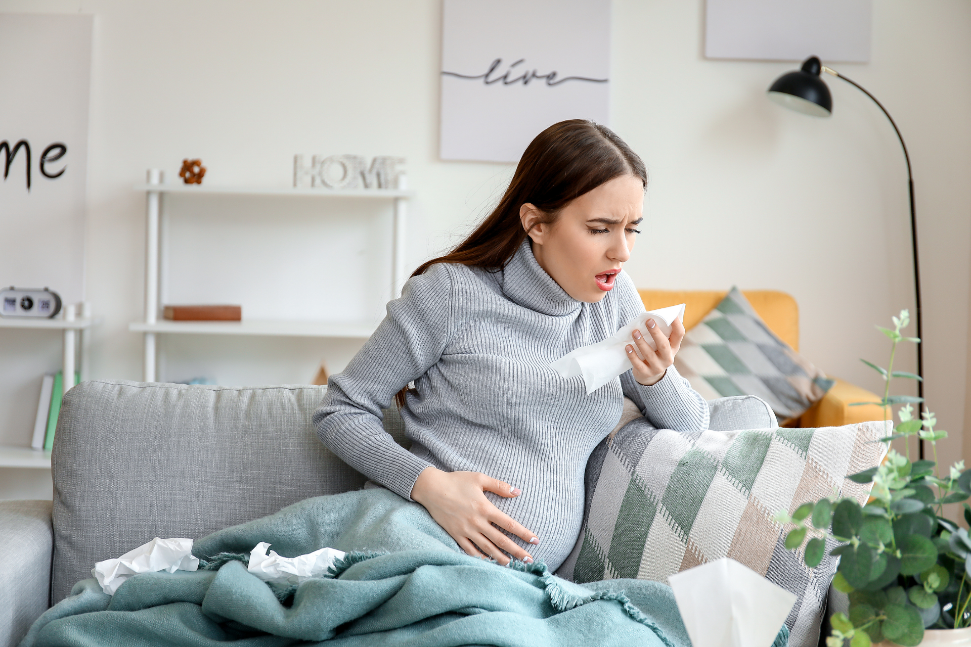 Pregnant woman sitting on a couch, coughing into a tissue while holding her stomach