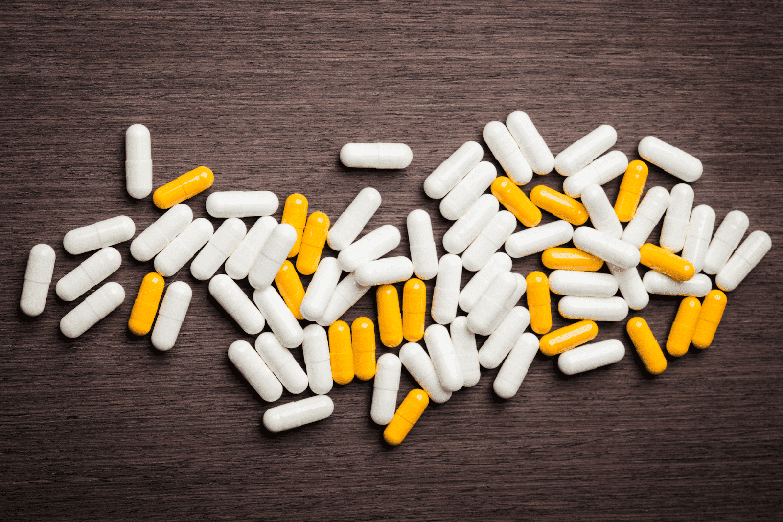 Scattered white and yellow capsules spread across a dark wooden surface, viewed from above, suggesting medication or dietary supplements.