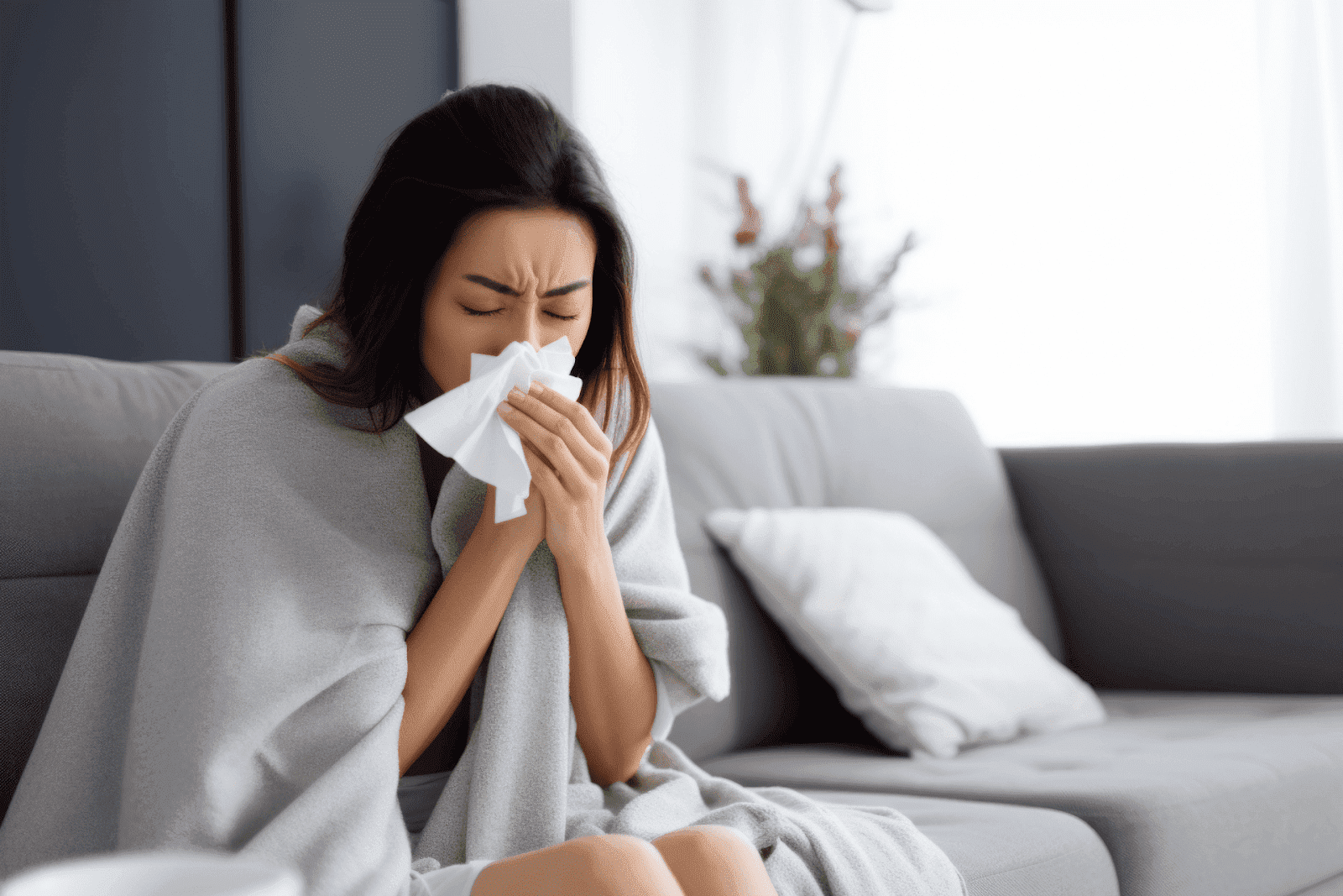 Sick woman wrapped in a blanket, blowing into a tissue while sitting on a couch.