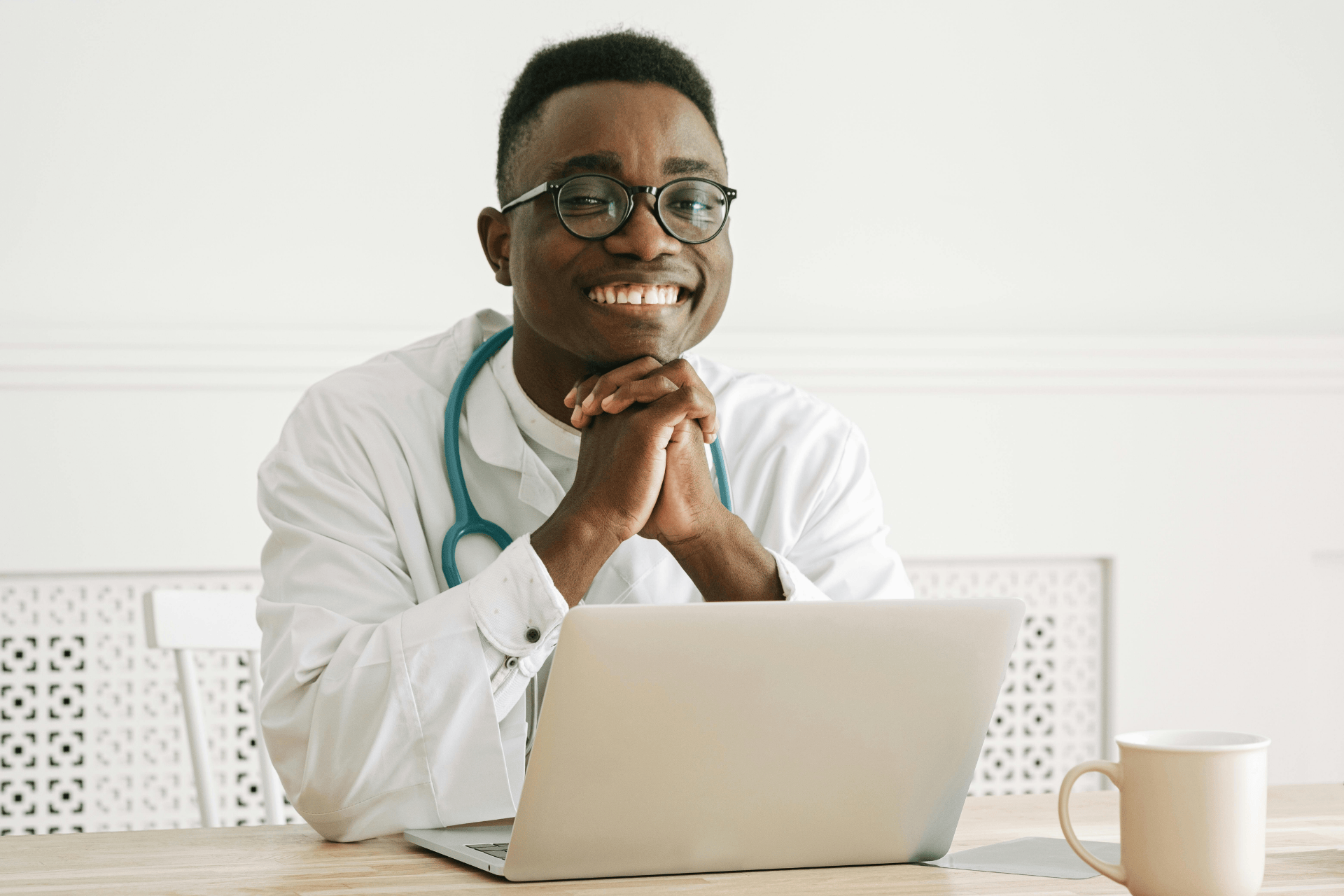 Smiling doctor with a stethoscope sitting at a desk with a laptop and a coffee cup.