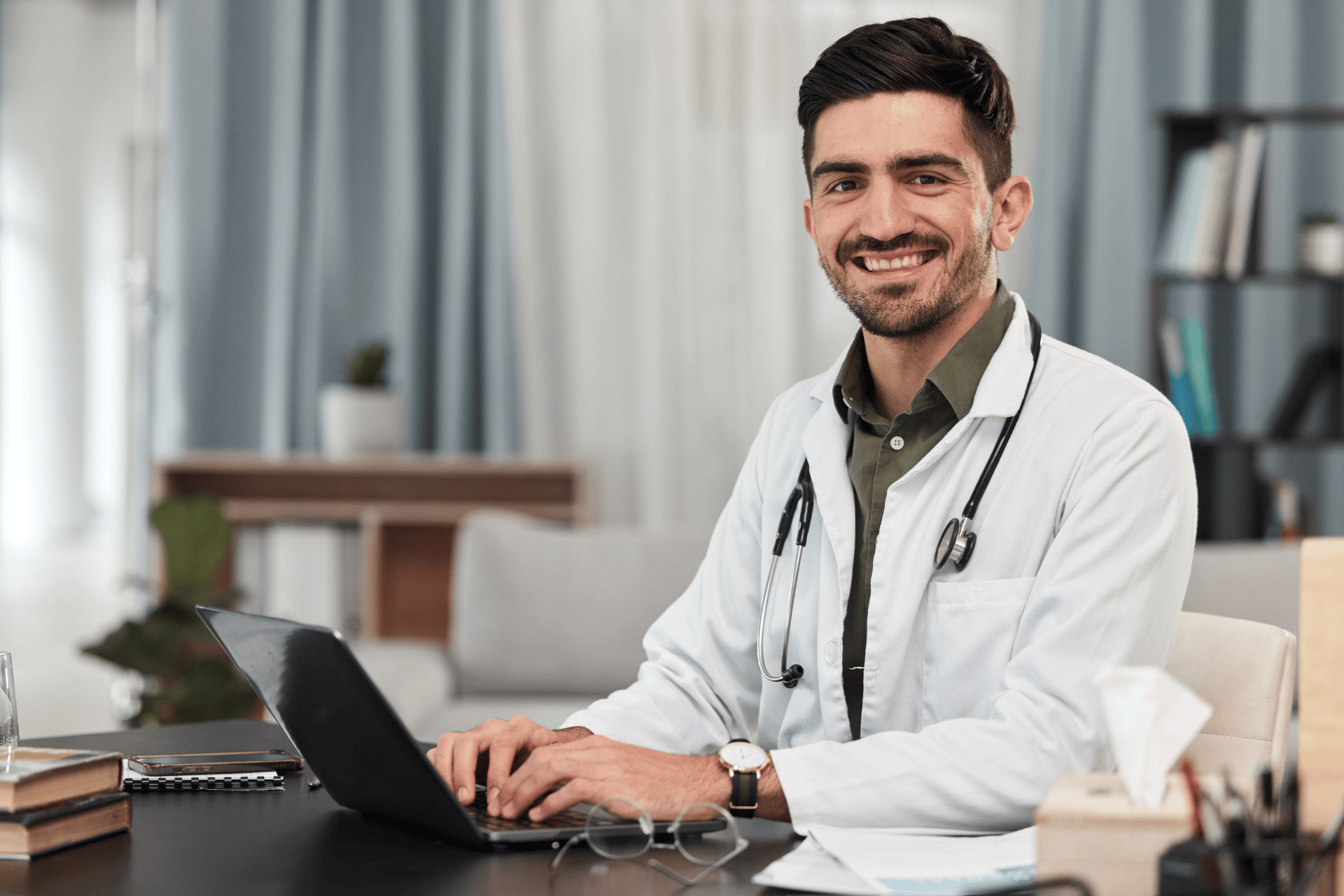 Smiling male doctor sitting at a desk typing on a laptop.