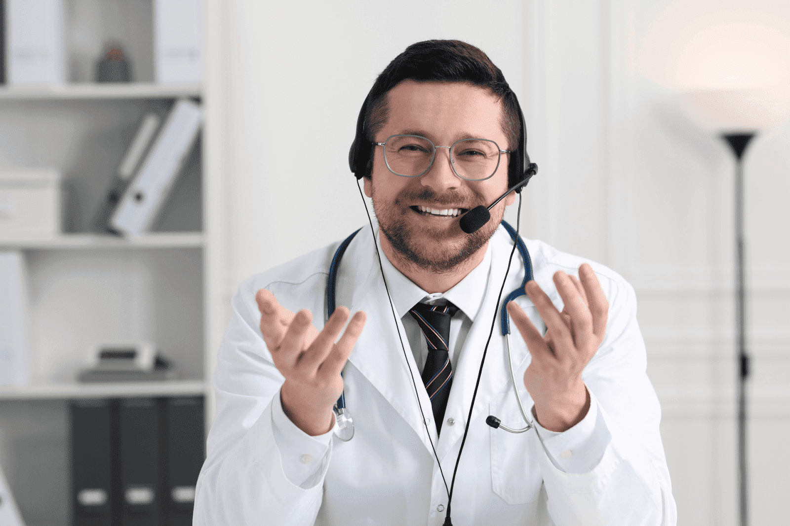 Smiling male doctor wearing a headset and stethoscope, speaking during a video call in an office.