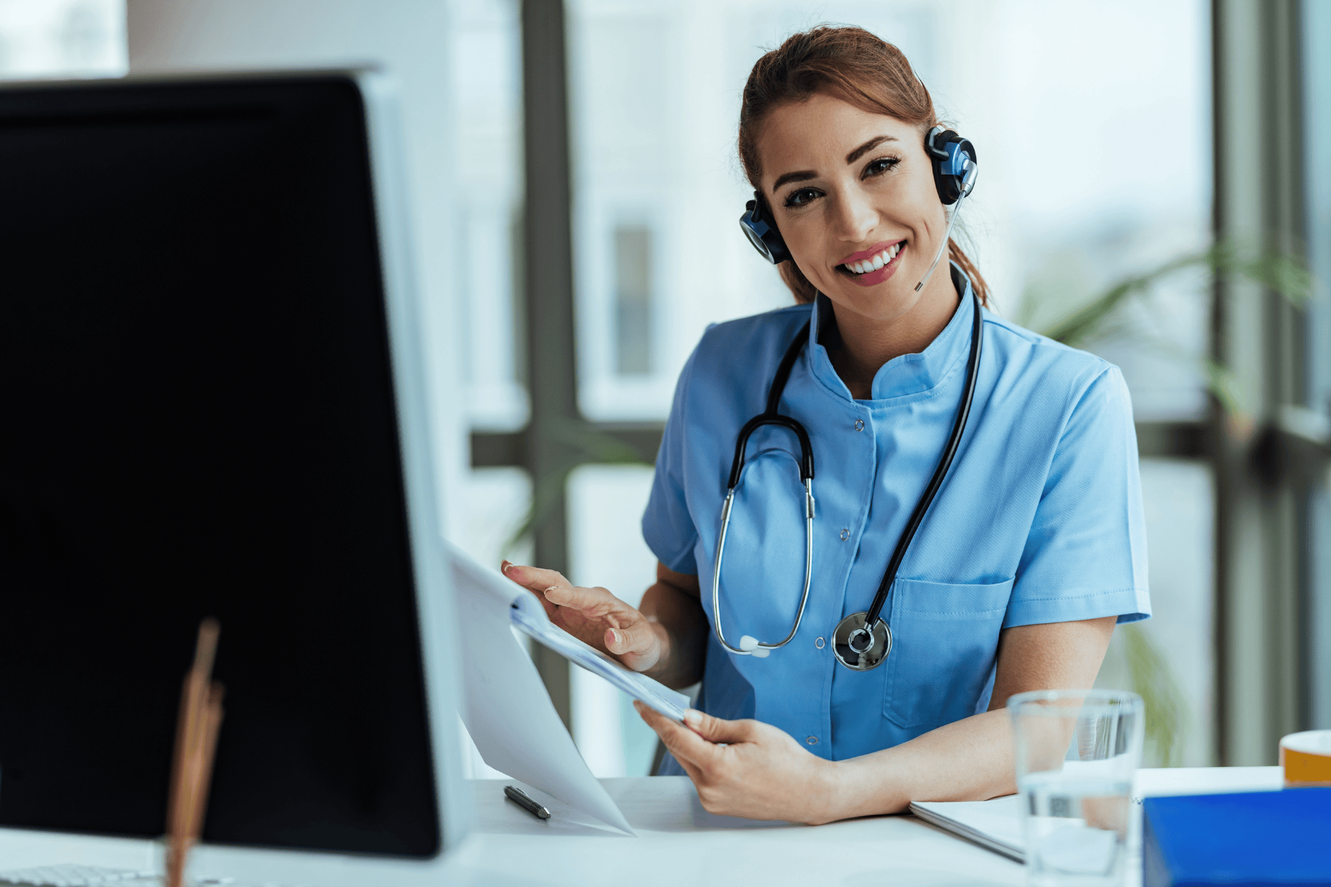Smiling nurse wearing a headset reviews documents at her desk.