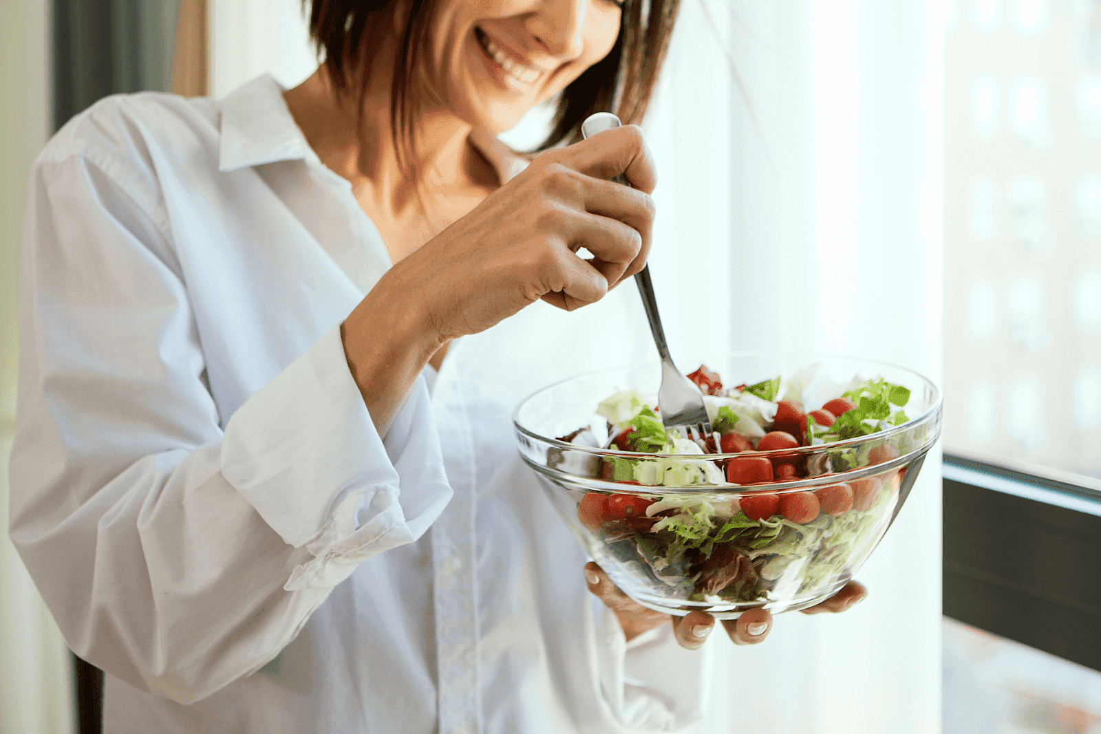 Smiling woman eating a fresh salad from a glass bowl.