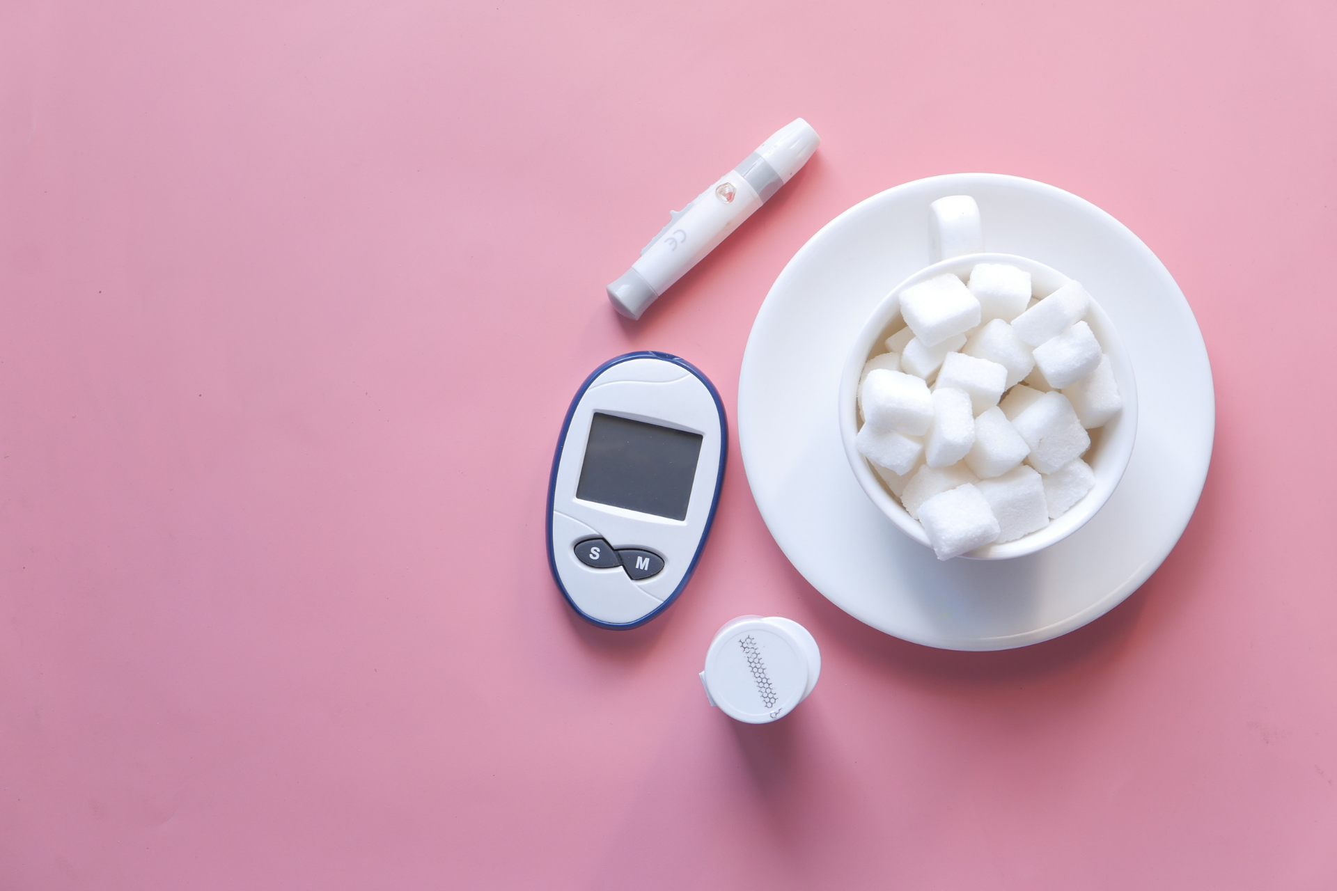 A glucose meter, lancing device, test-strip vial, and a cup of sugar cubes arranged on a pink background
