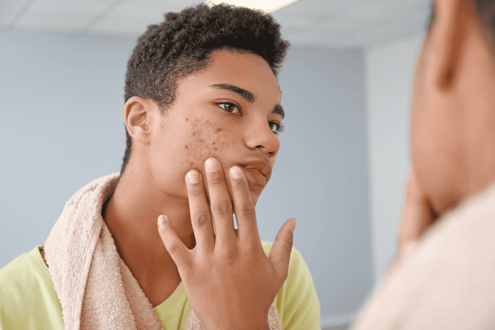 Teen boy with a towel around his neck, examining acne on his cheek in the mirror