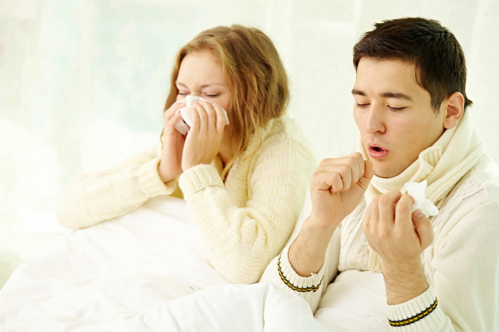 Two people sitting in bed wearing sweaters, coughing and blowing their noses with tissues, appearing sick