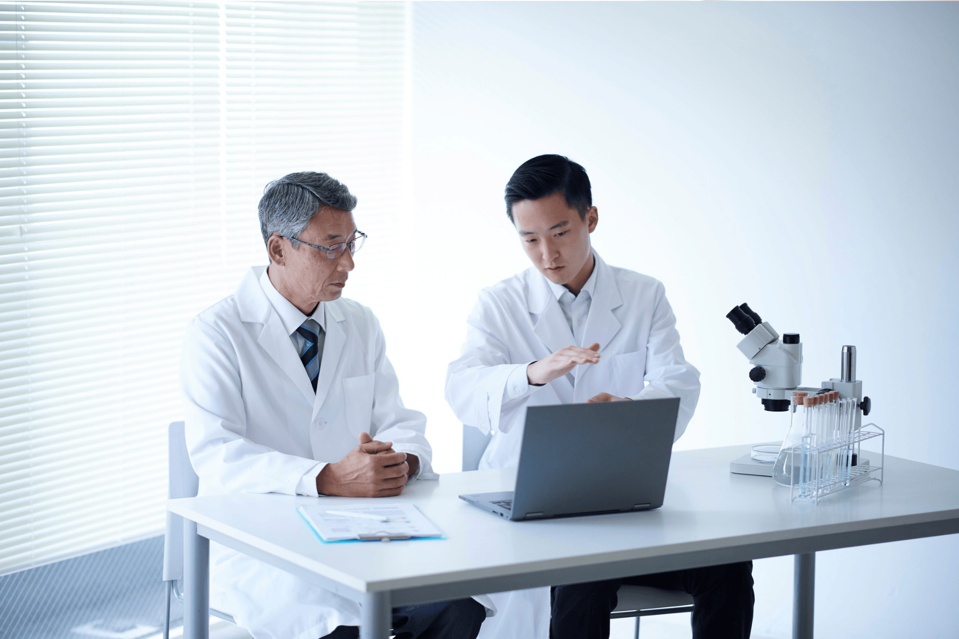 Two doctors in lab coats discussing research data on a laptop beside lab equipment.