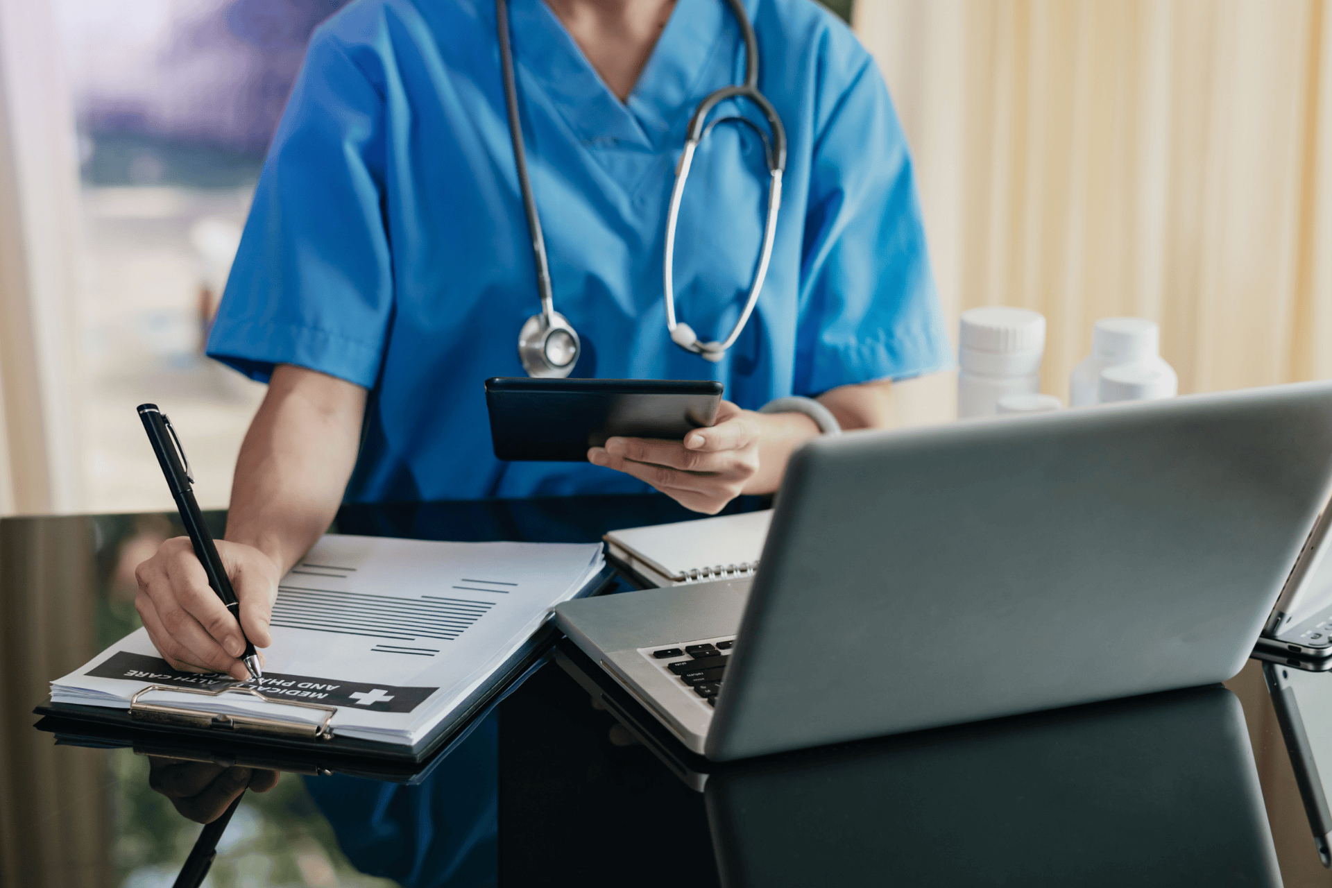 Healthcare professional in blue scrubs working on documents and a tablet beside a laptop.