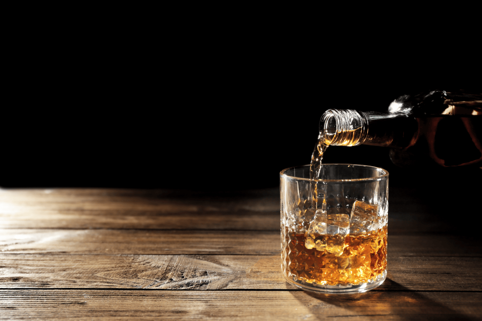 Whiskey being poured into a glass with ice cubes on a wooden table.