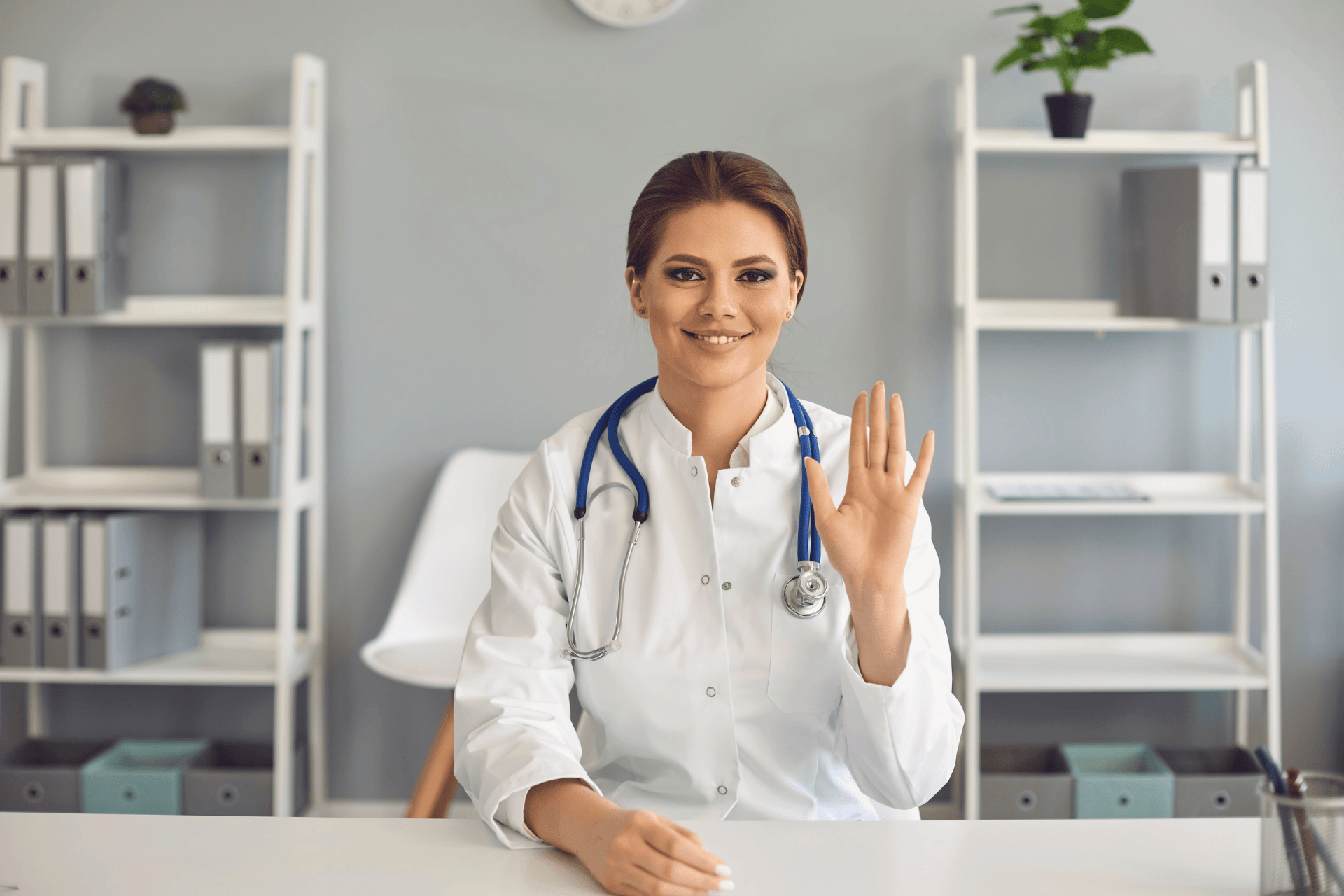 Smiling female doctor with a stethoscope waving while sitting at a desk.