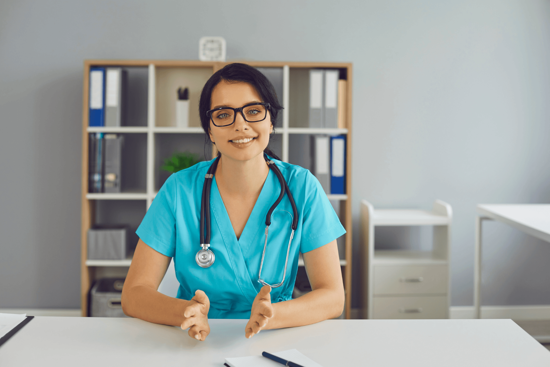 Female doctor in scrubs sitting at a desk and speaking during a consultation.