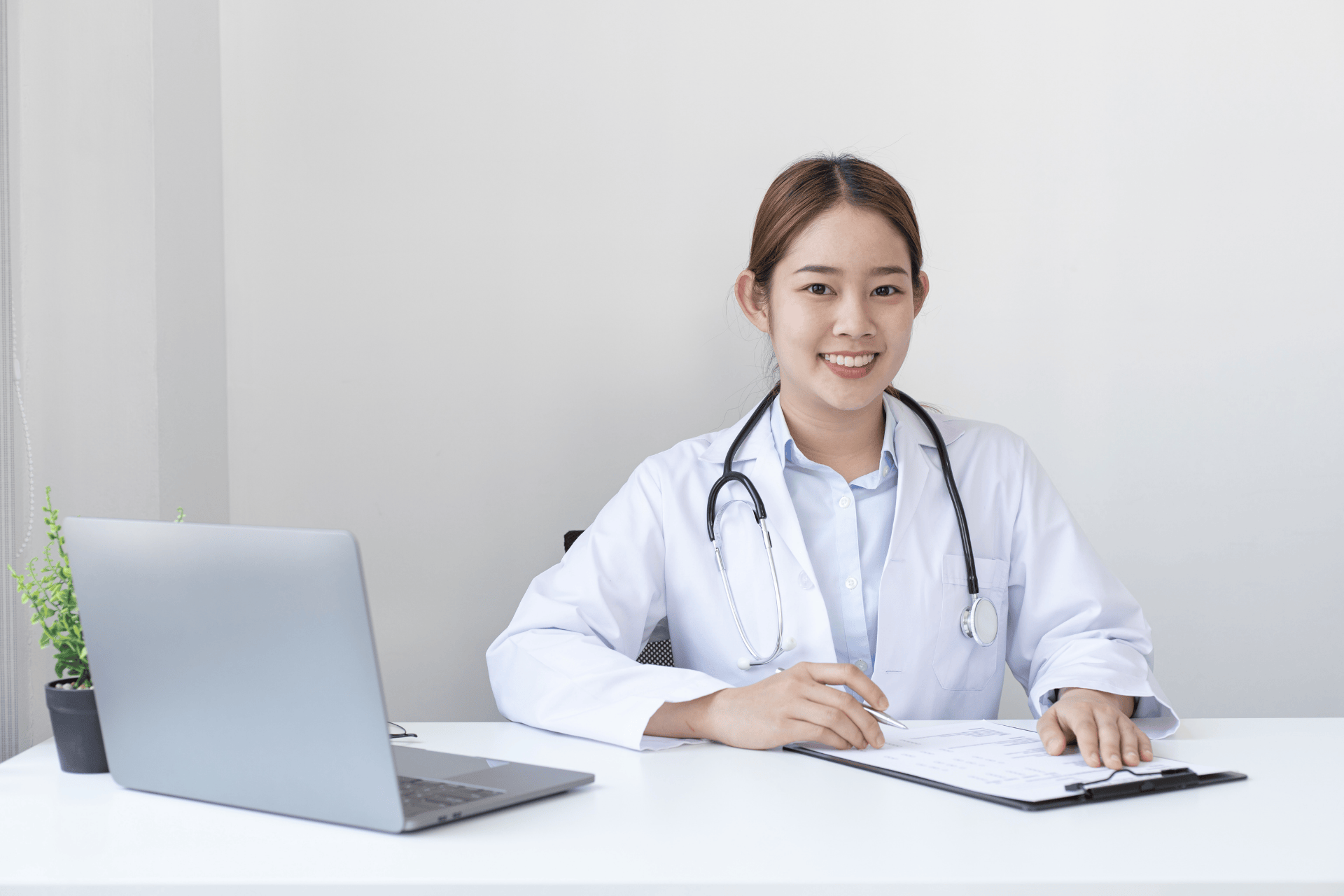 Female doctor sitting at a desk, smiling while writing on a clipboard beside a laptop.