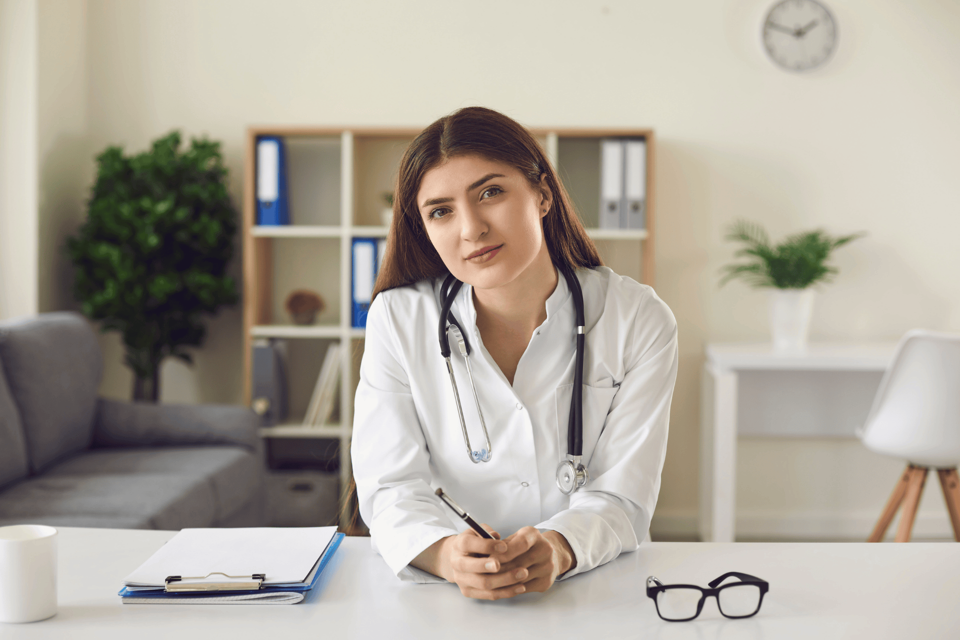 Female doctor sitting at a desk, holding a pen and looking forward.