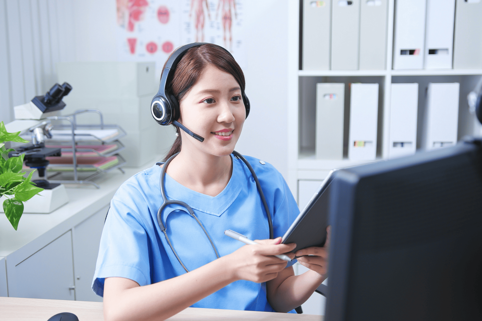 Doctor wearing a headset and scrubs, holding a tablet while speaking during a telehealth session.