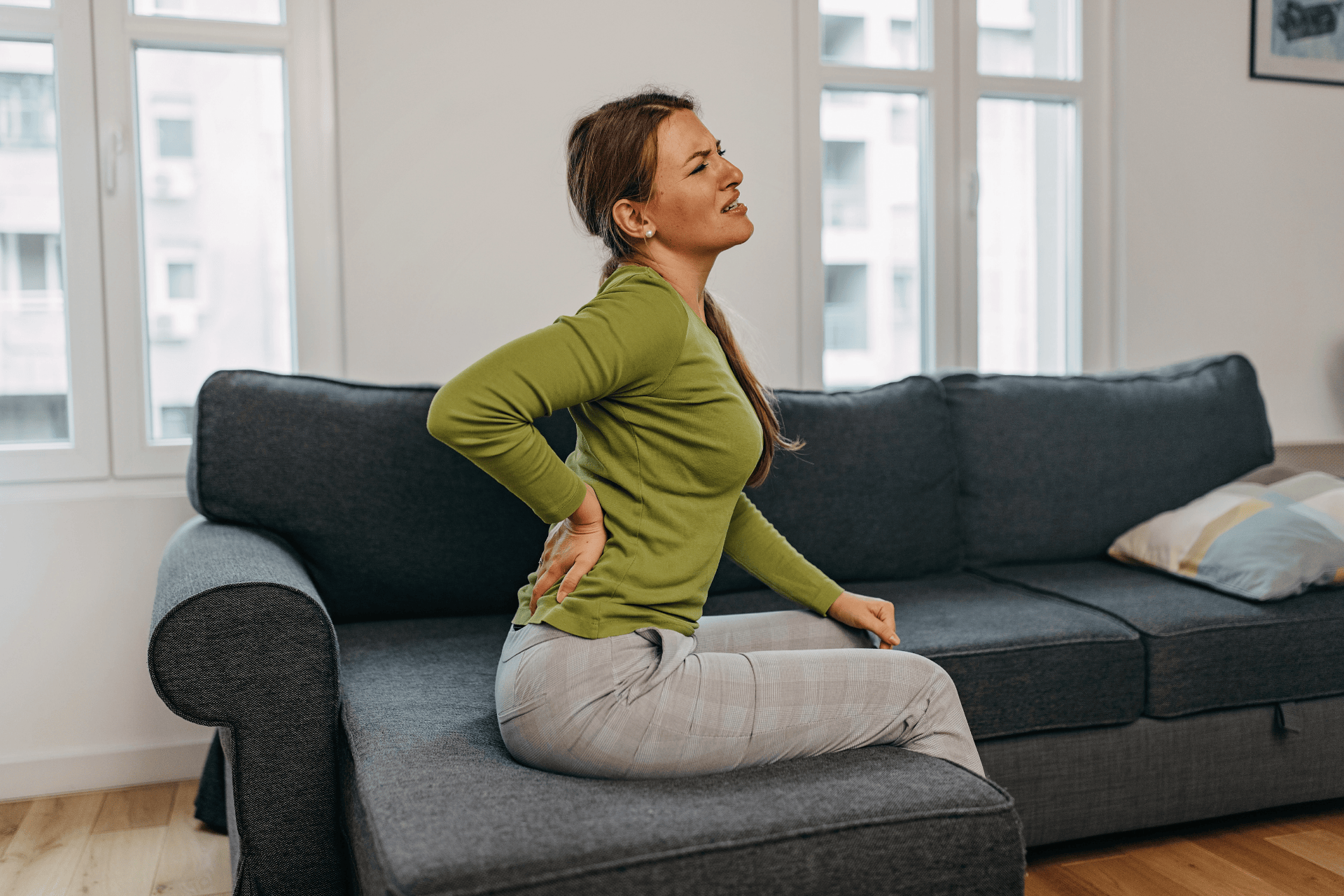 Young woman sitting on a couch holding her lower back in pain.
