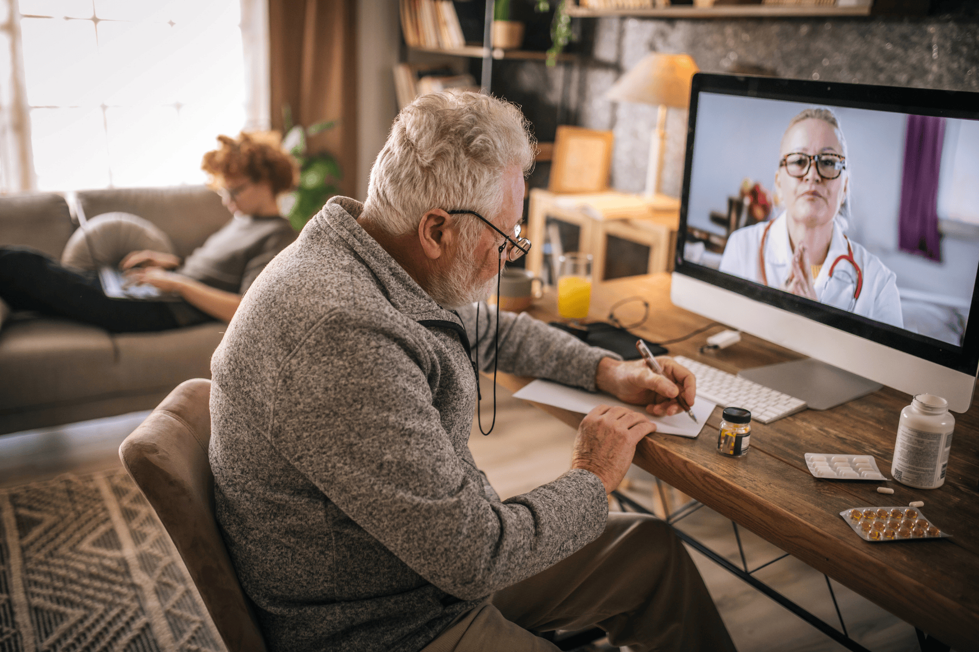 An older man on a video call with a doctor while writing notes at his desk.