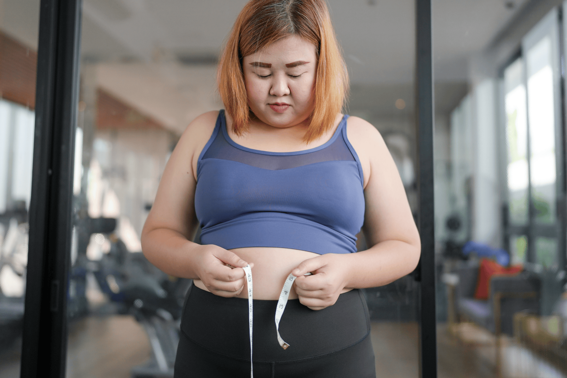 Woman in workout clothes measuring her waist with a tape measure in a gym.