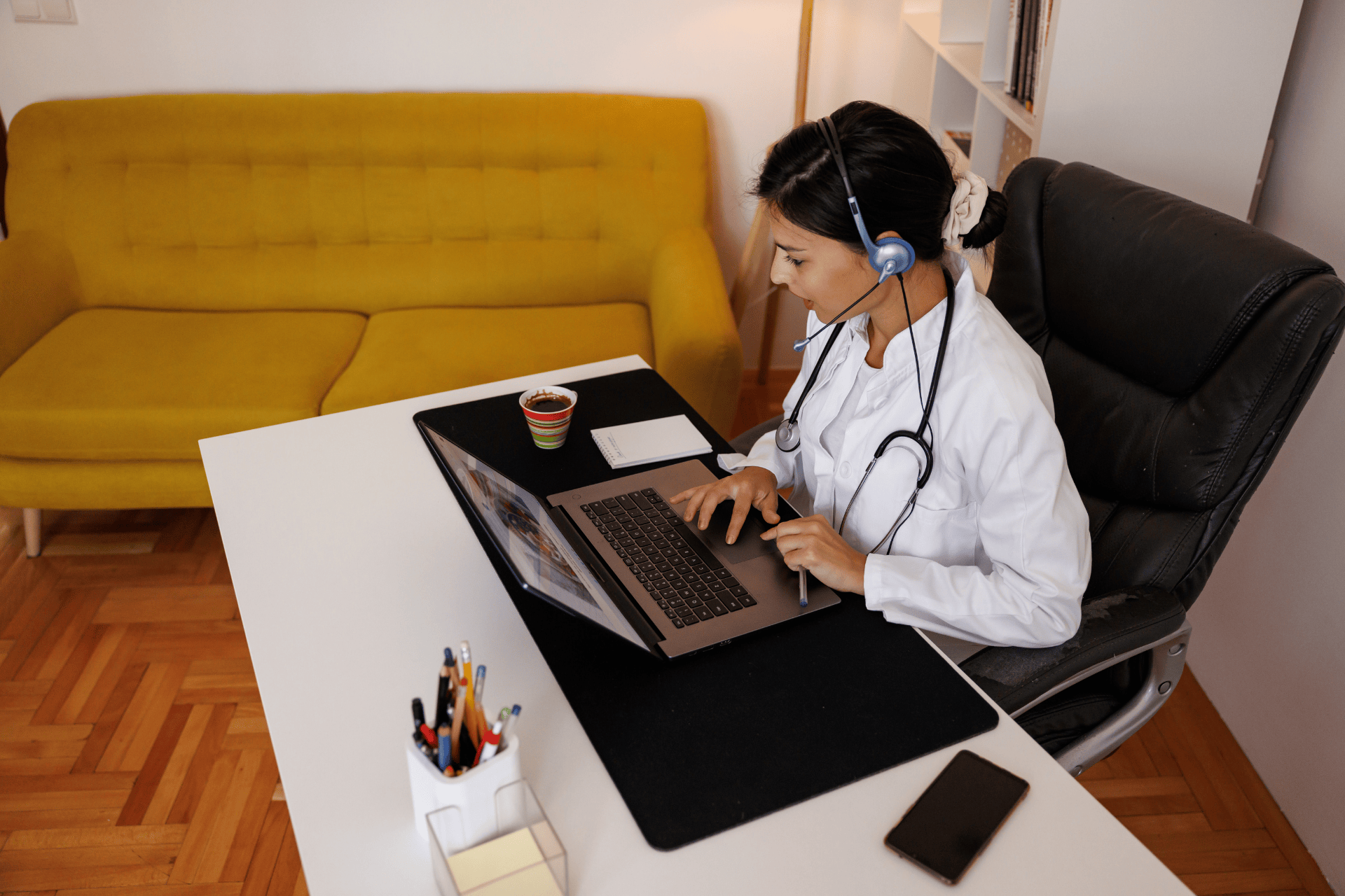 Doctor wearing a headset works on a laptop while taking notes.