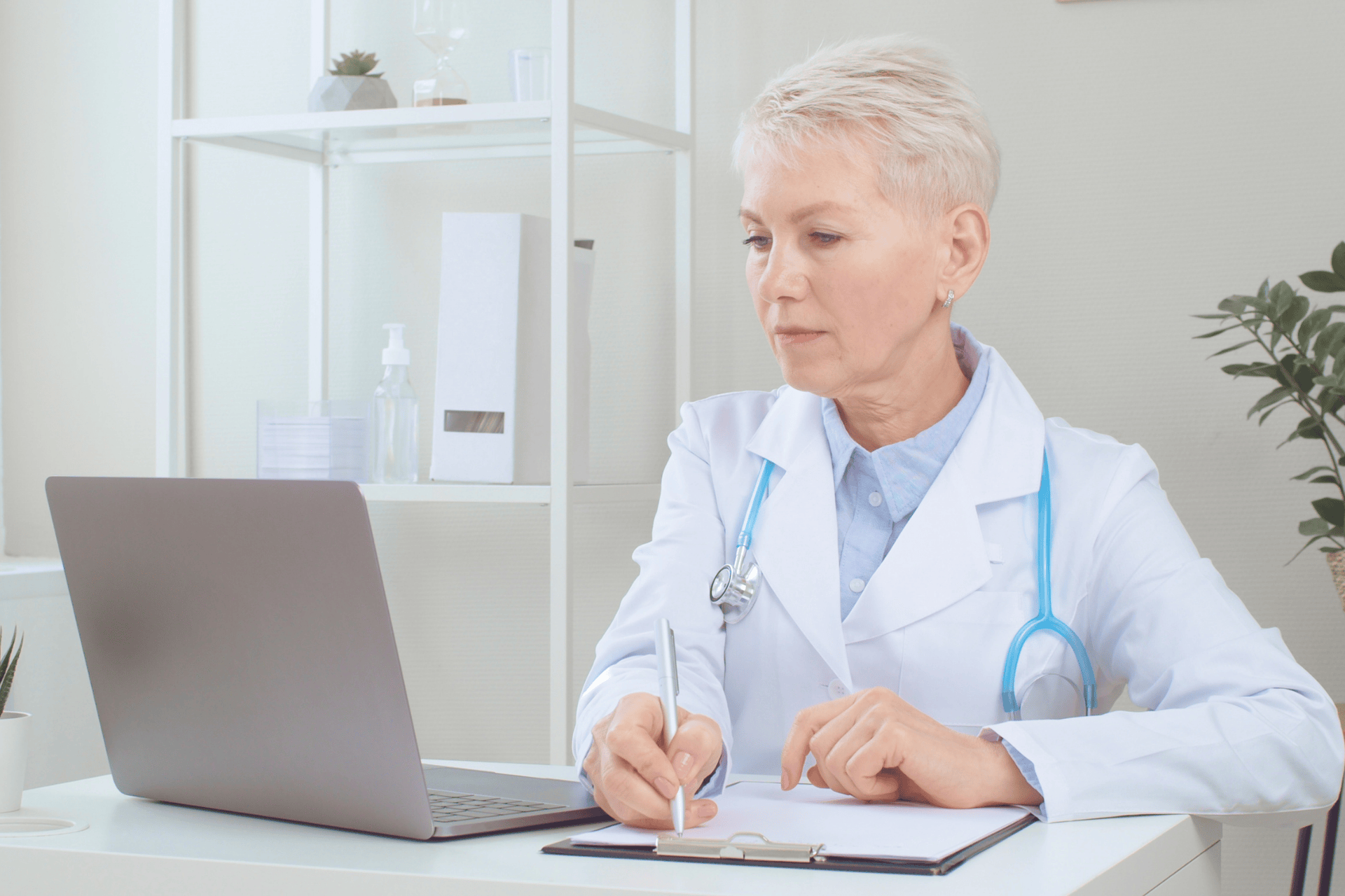 Older female doctor writing on a clipboard while looking at a laptop.