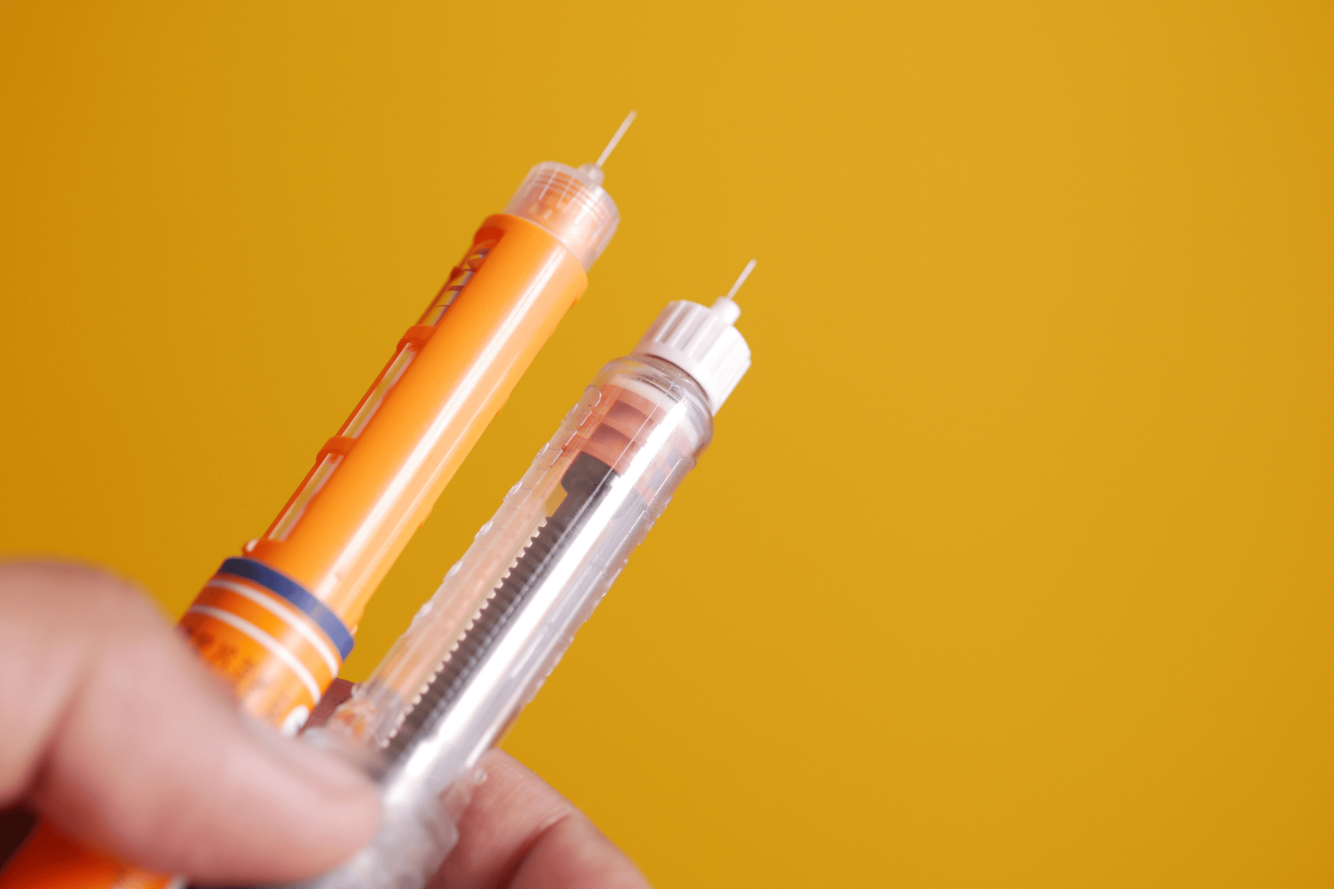 Hand holding two insulin pens with needles against a yellow background