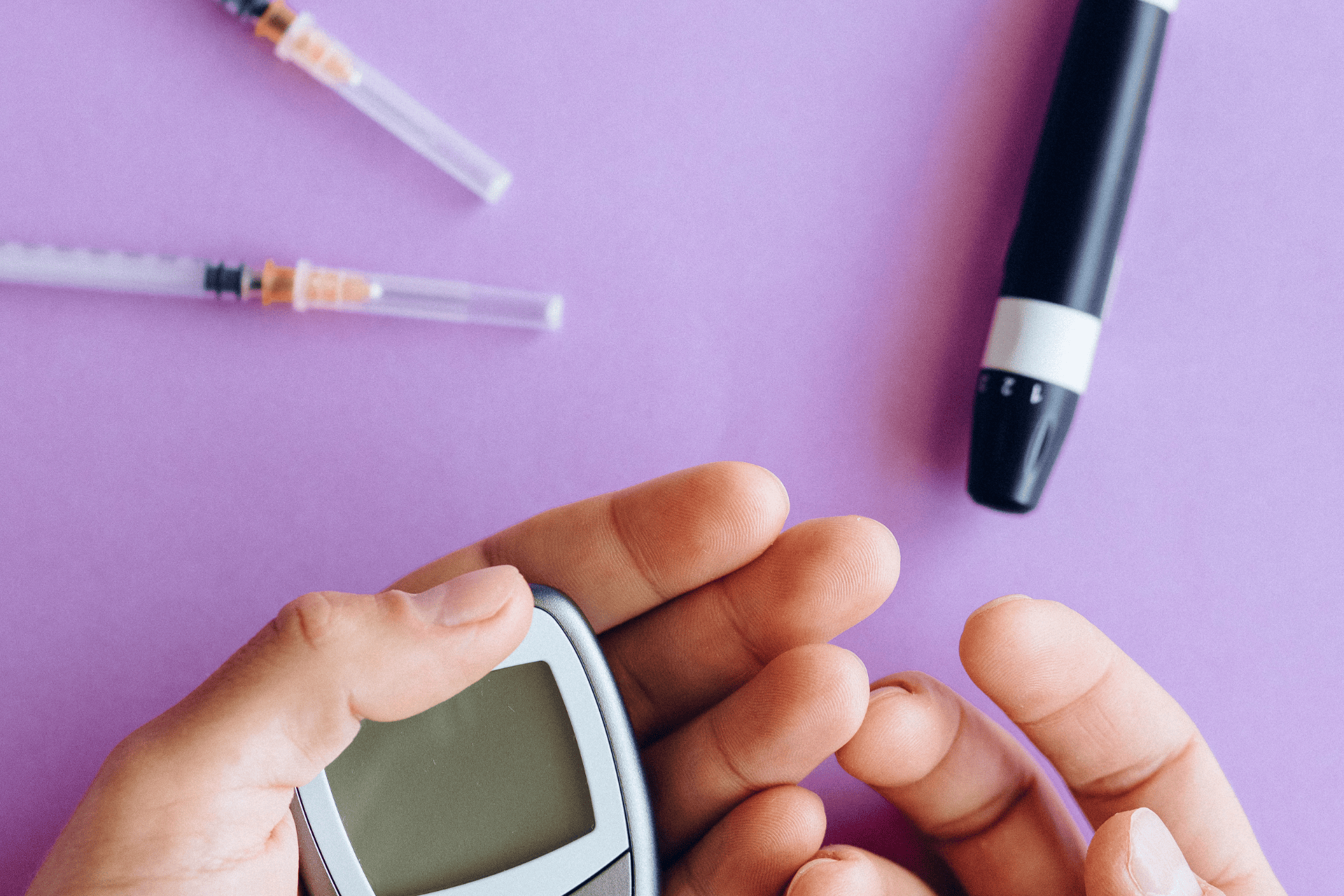 Close-up of a person checking their blood sugar with a glucose meter, syringes and a lancet device nearby.