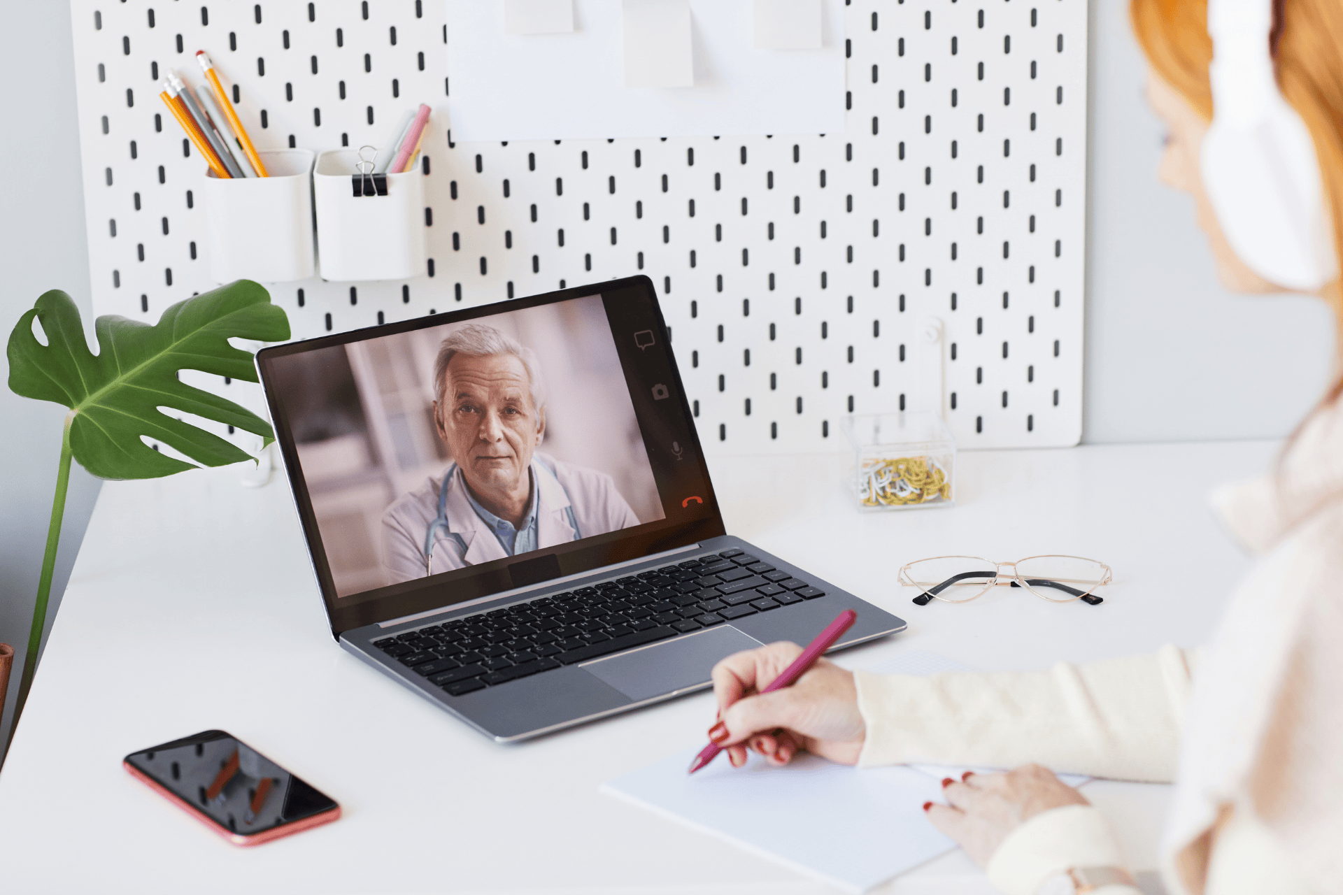 Woman taking notes during an online video call with a doctor displayed on a laptop screen.