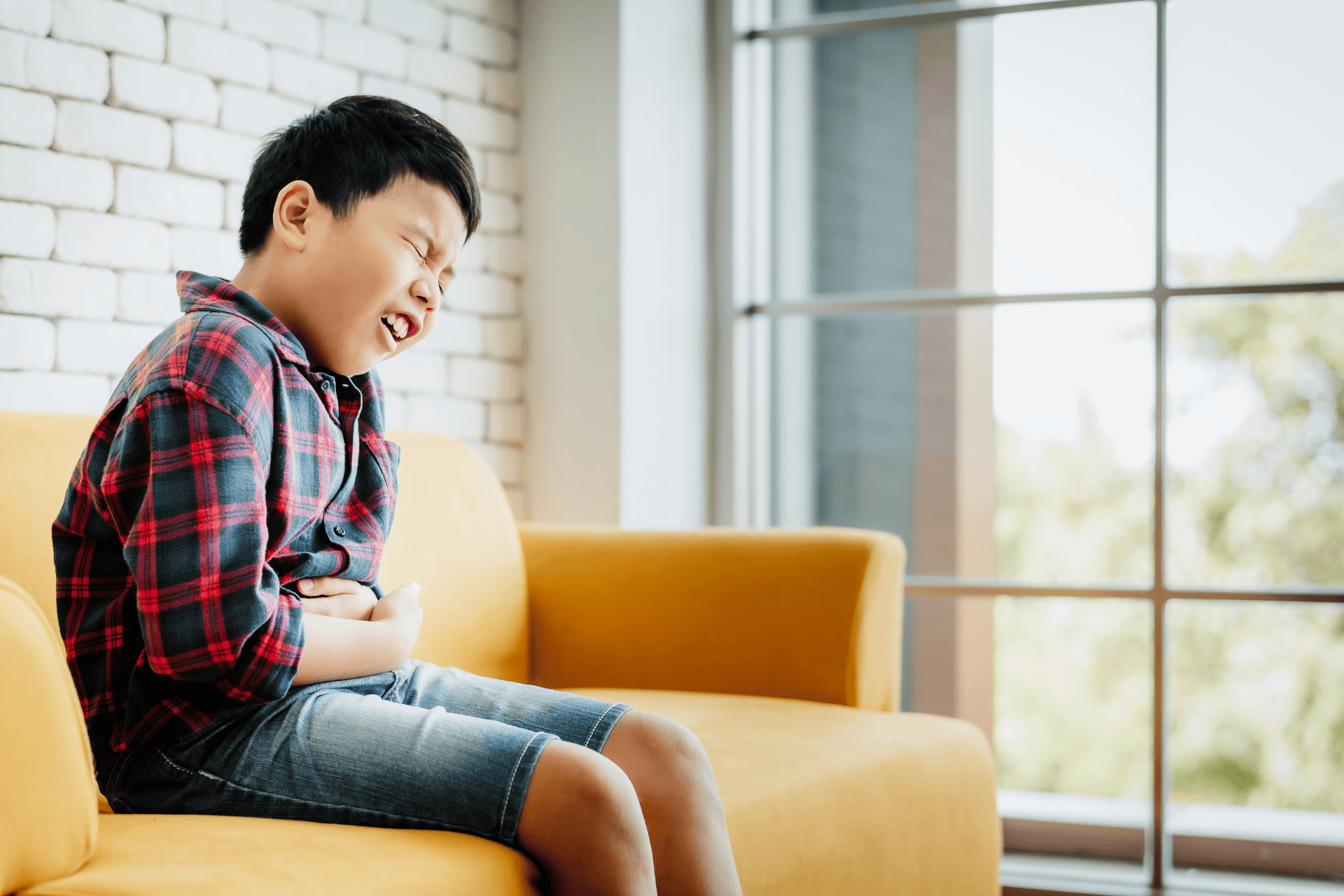 Young boy sitting on a couch holding his lower abdomen in pain.