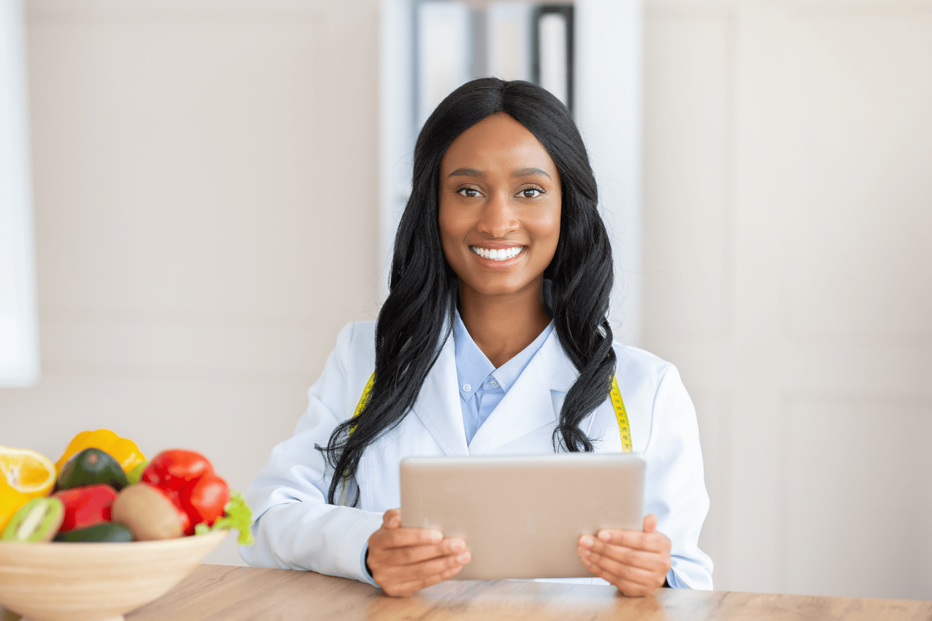 Smiling female doctor in a white coat holding a tablet, with fresh fruits and vegetables on the table.